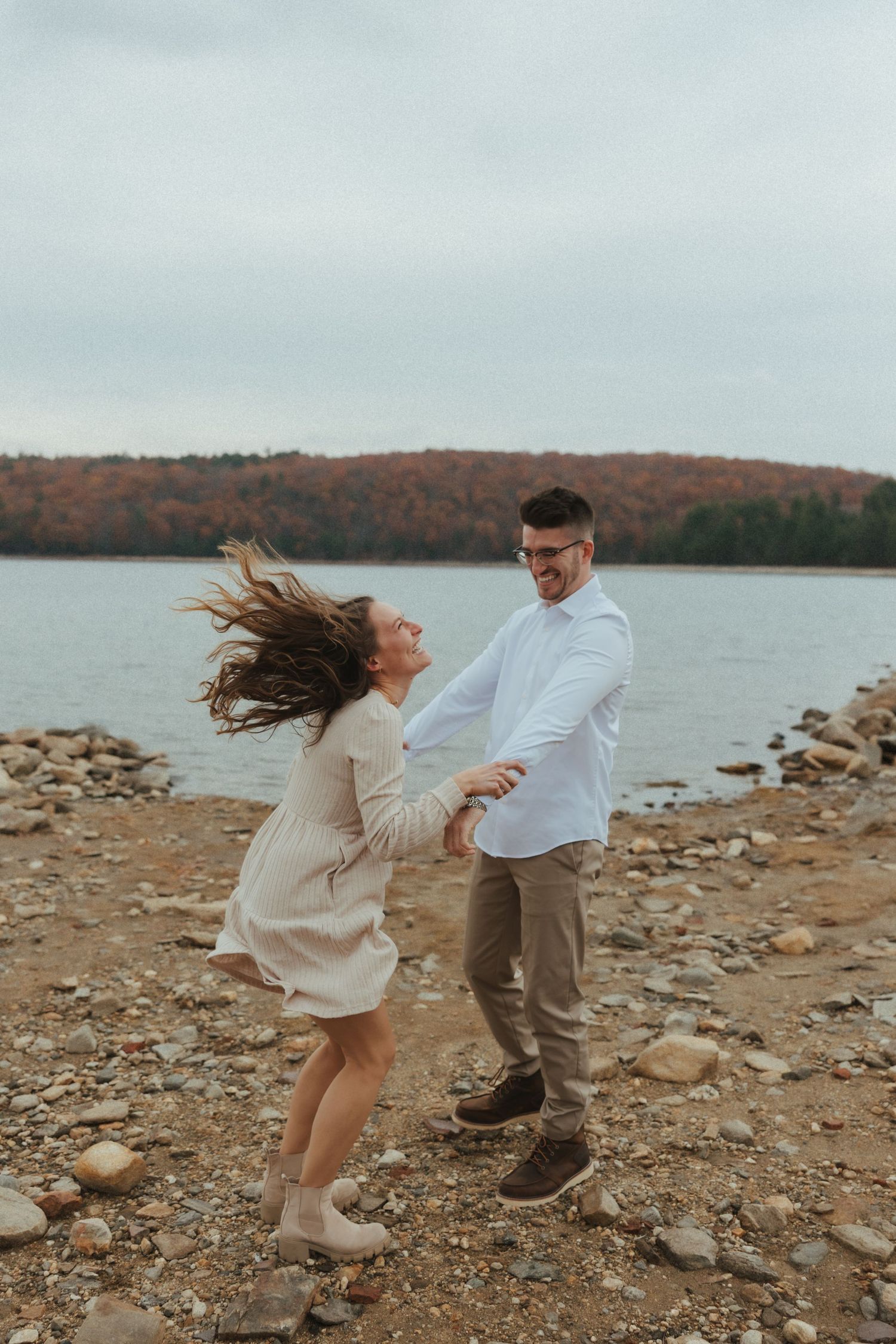A couple dances playfully on a rocky shoreline with autumn trees and a lake in the background.