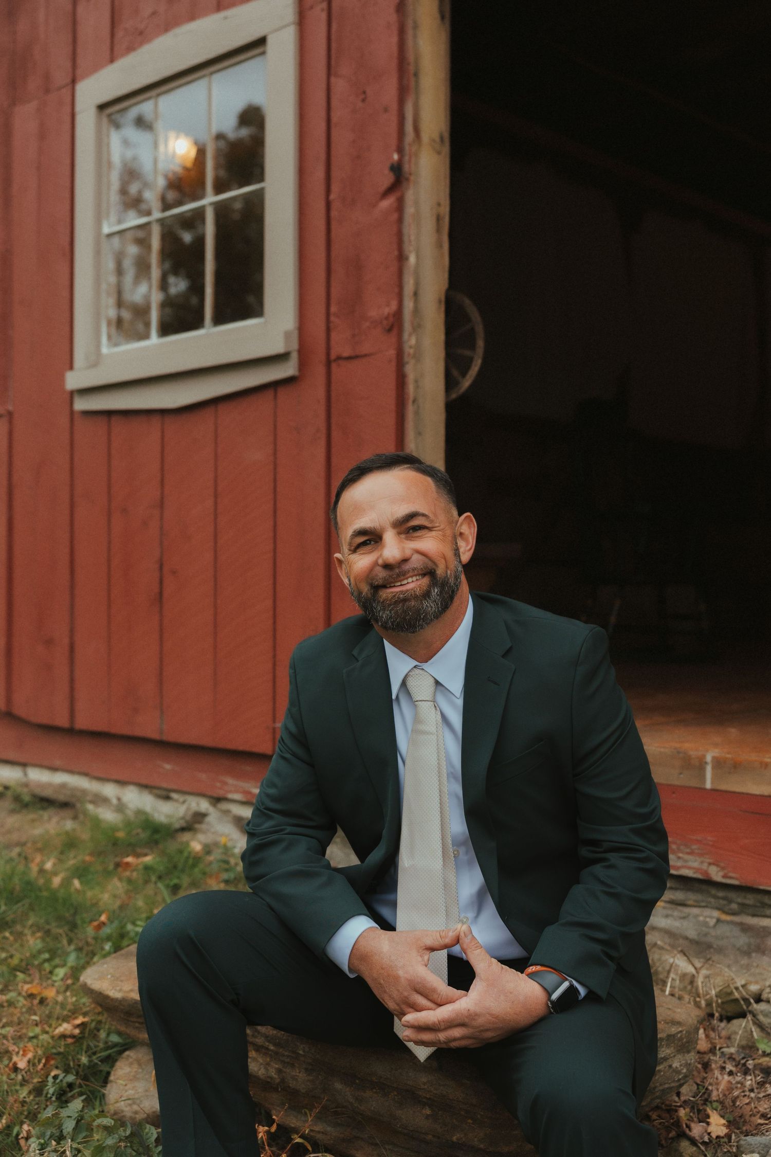 Professional portrait of someone in a dark suit sitting outside a rustic red barn with white-trimmed windows.