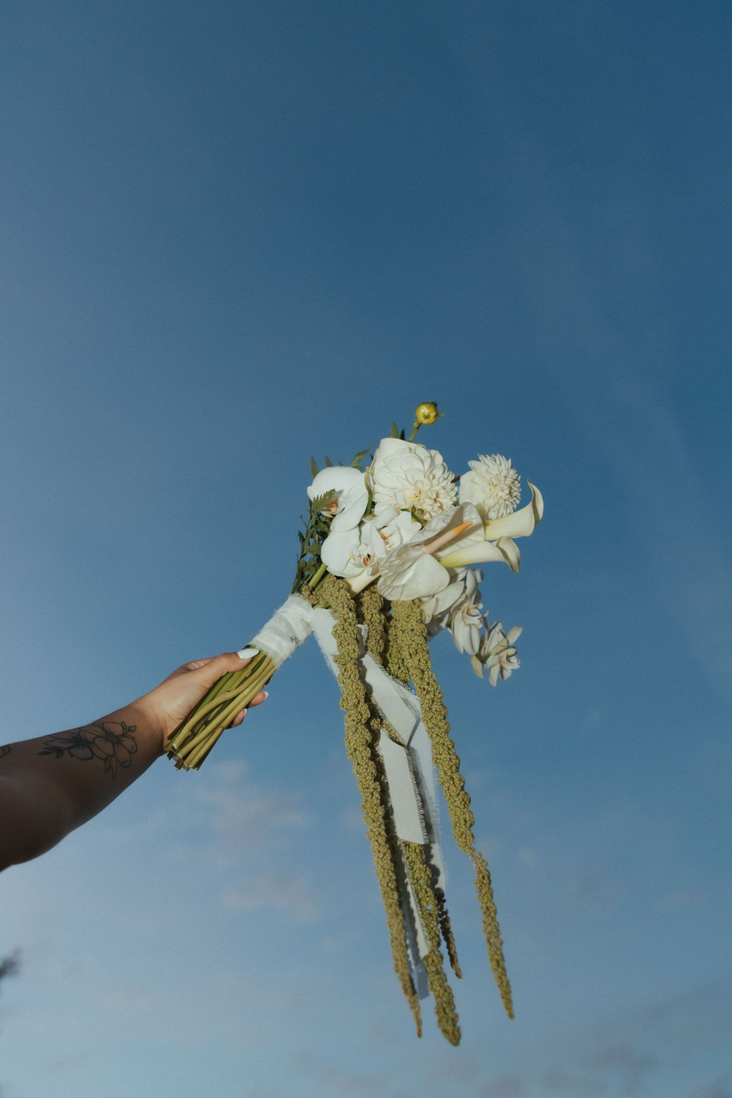 Hand holding fresh white asparagus and yellow flowers against a vibrant blue sky background.