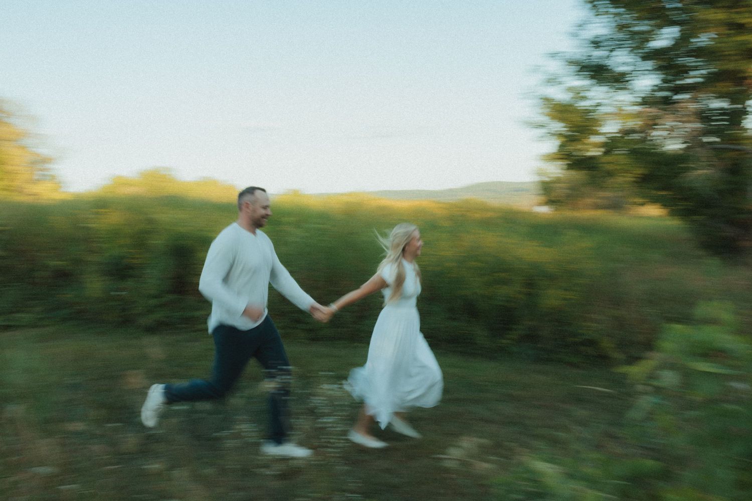 A couple in wedding attire runs playfully through a sunlit meadow at golden hour.
