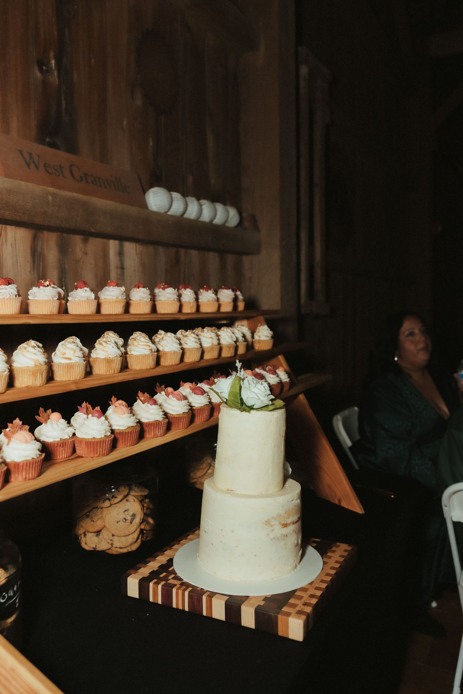 A white wedding cake displayed on a wooden stand with rows of cupcakes arranged on shelves behind it.