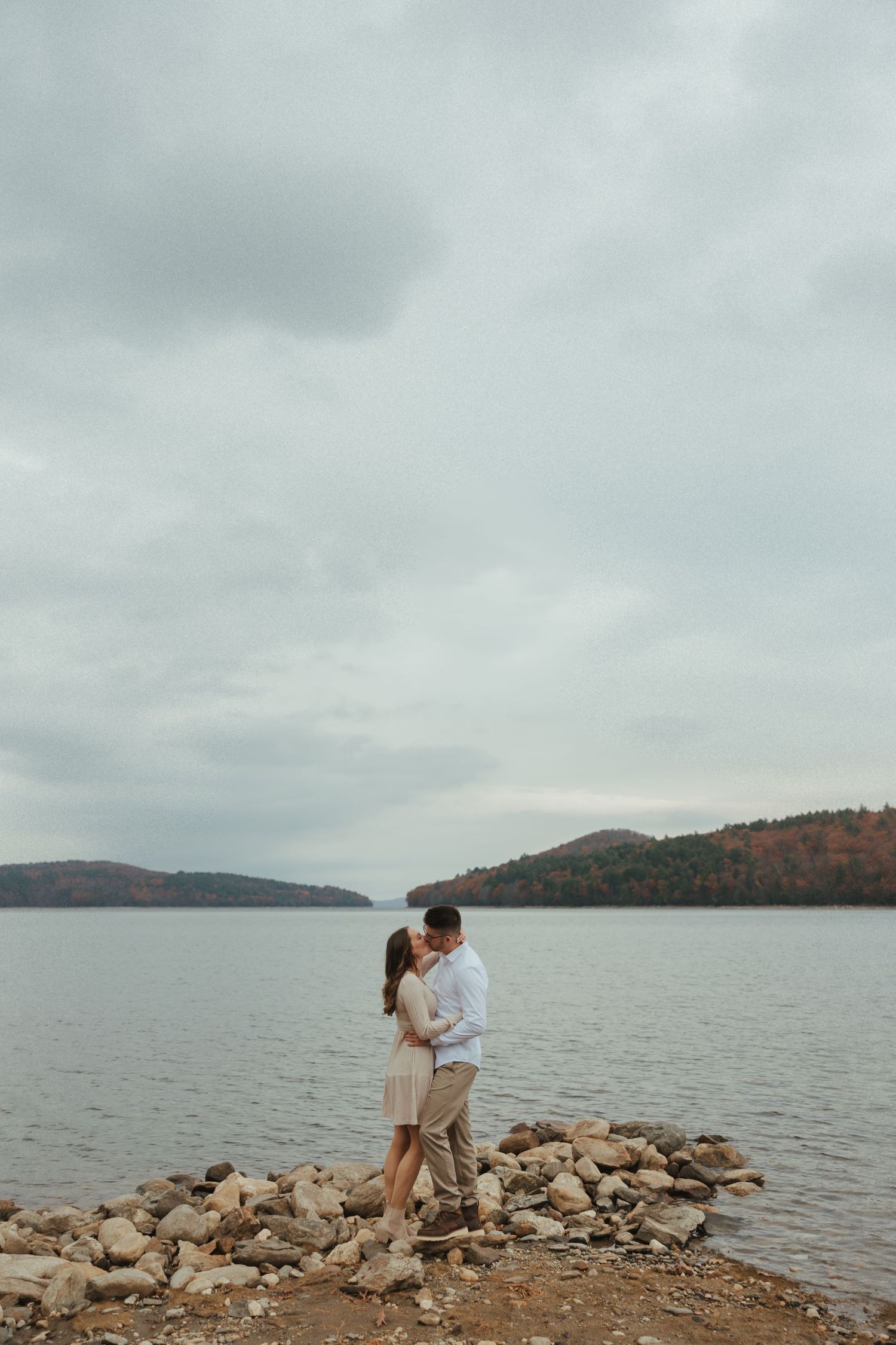 Couple embraces on rocky shoreline with misty lake and autumn mountains in background.