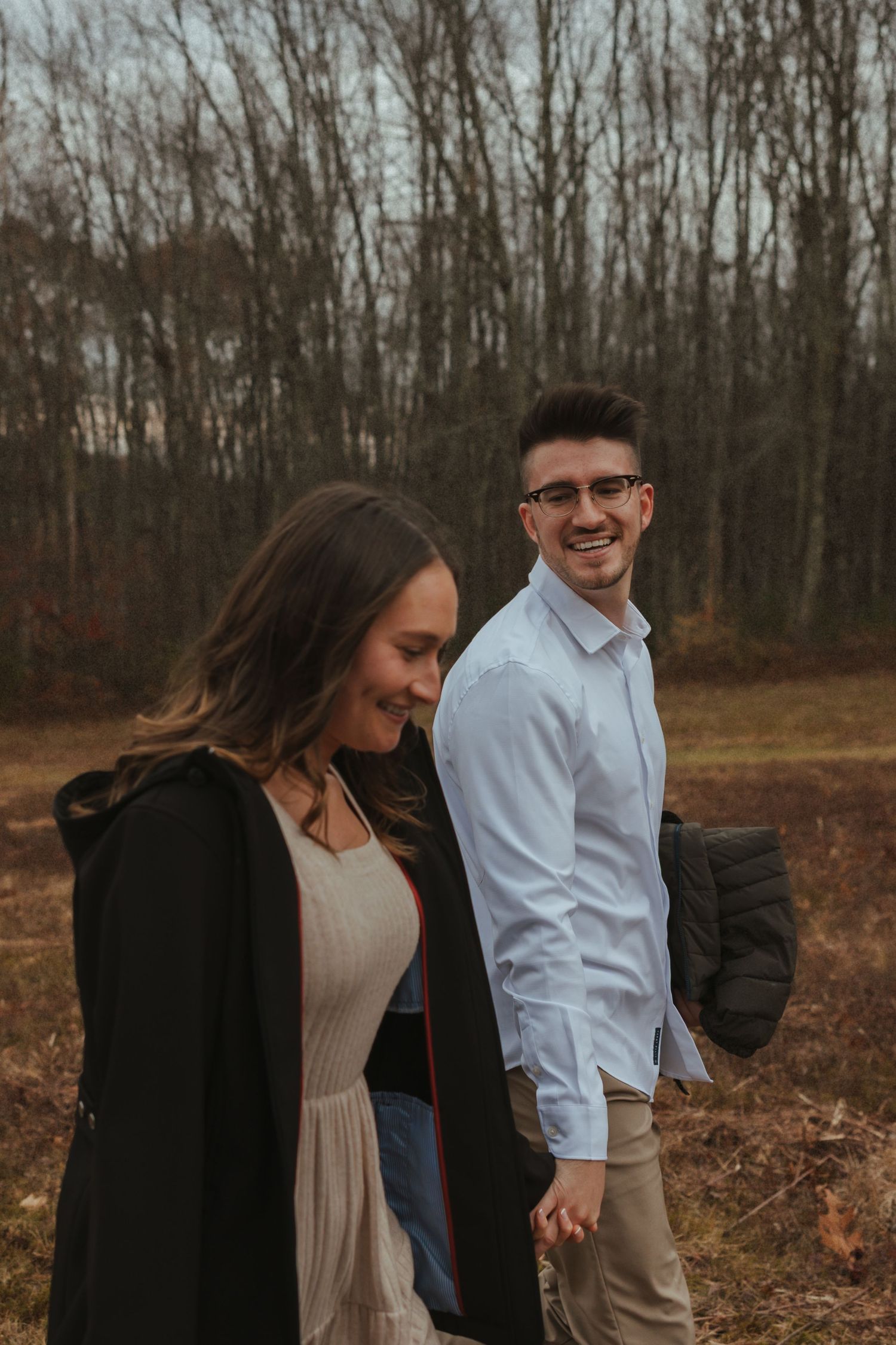 A couple walks together through a wooded area during an autumn engagement photo session.