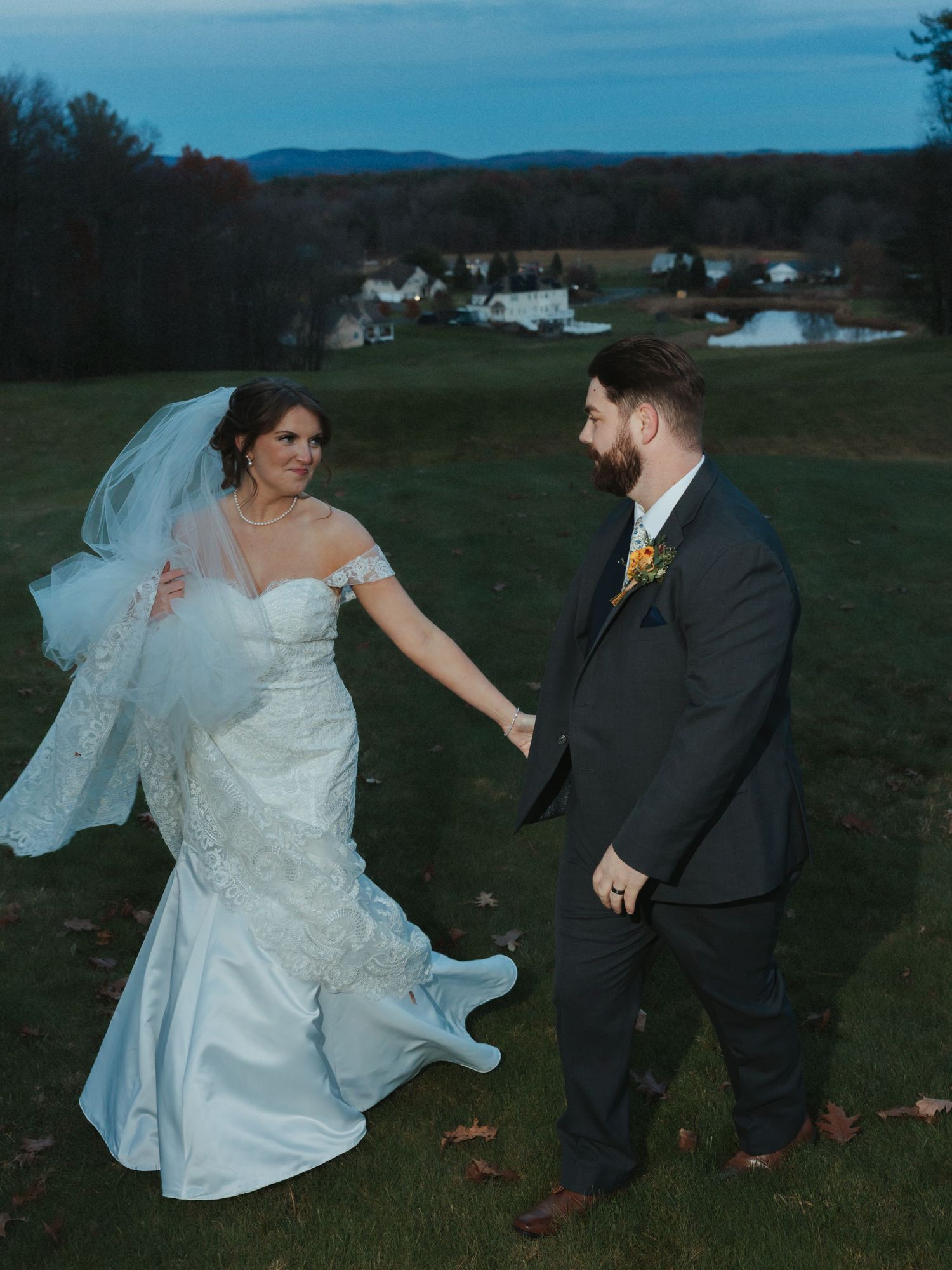 A couple in wedding attire share a romantic moment on a hilltop overlooking a rural landscape at dusk.
