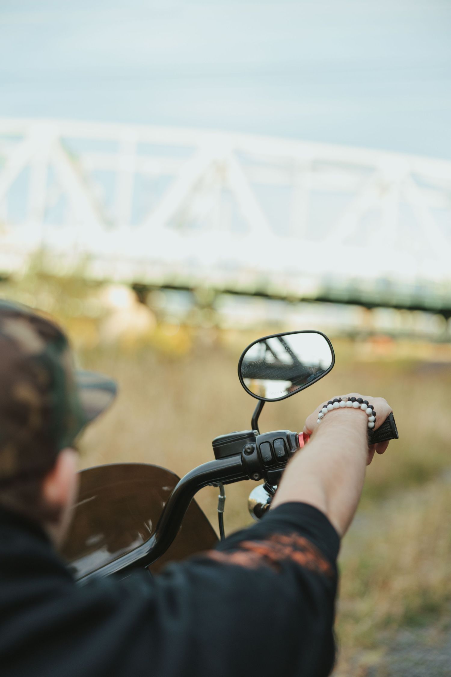 A motorcycle side mirror and handlebar shown in focus with a blurred bridge structure in the background.