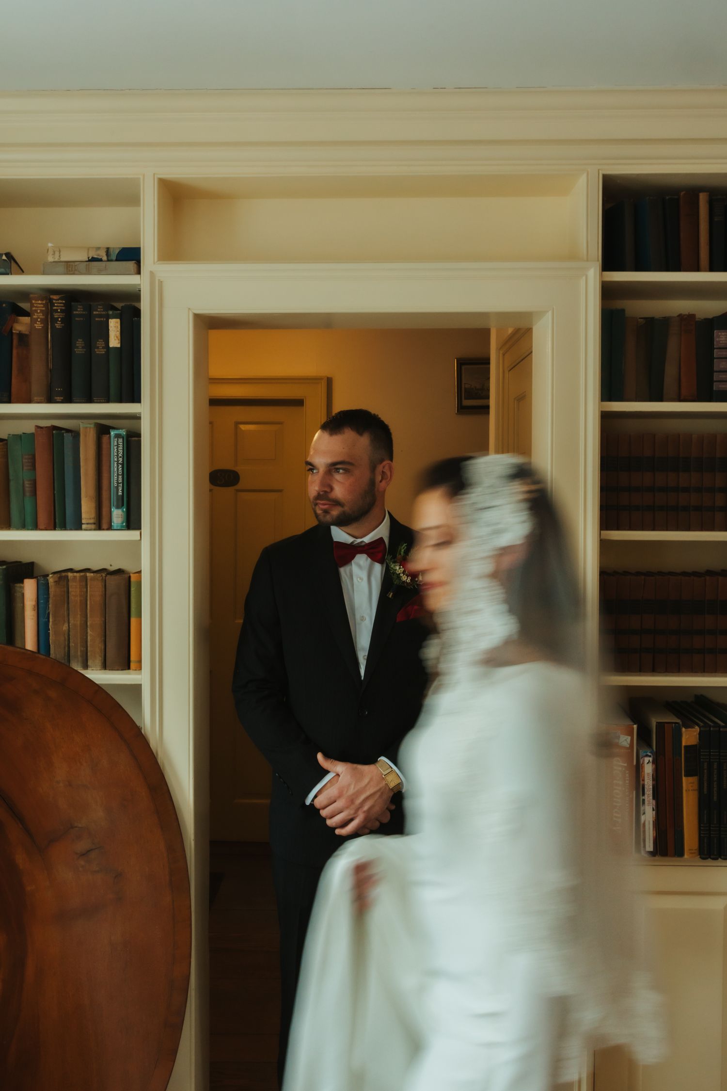 A wedding couple stands in a library room with built-in bookshelves, the bride in white and groom in black tie attire.