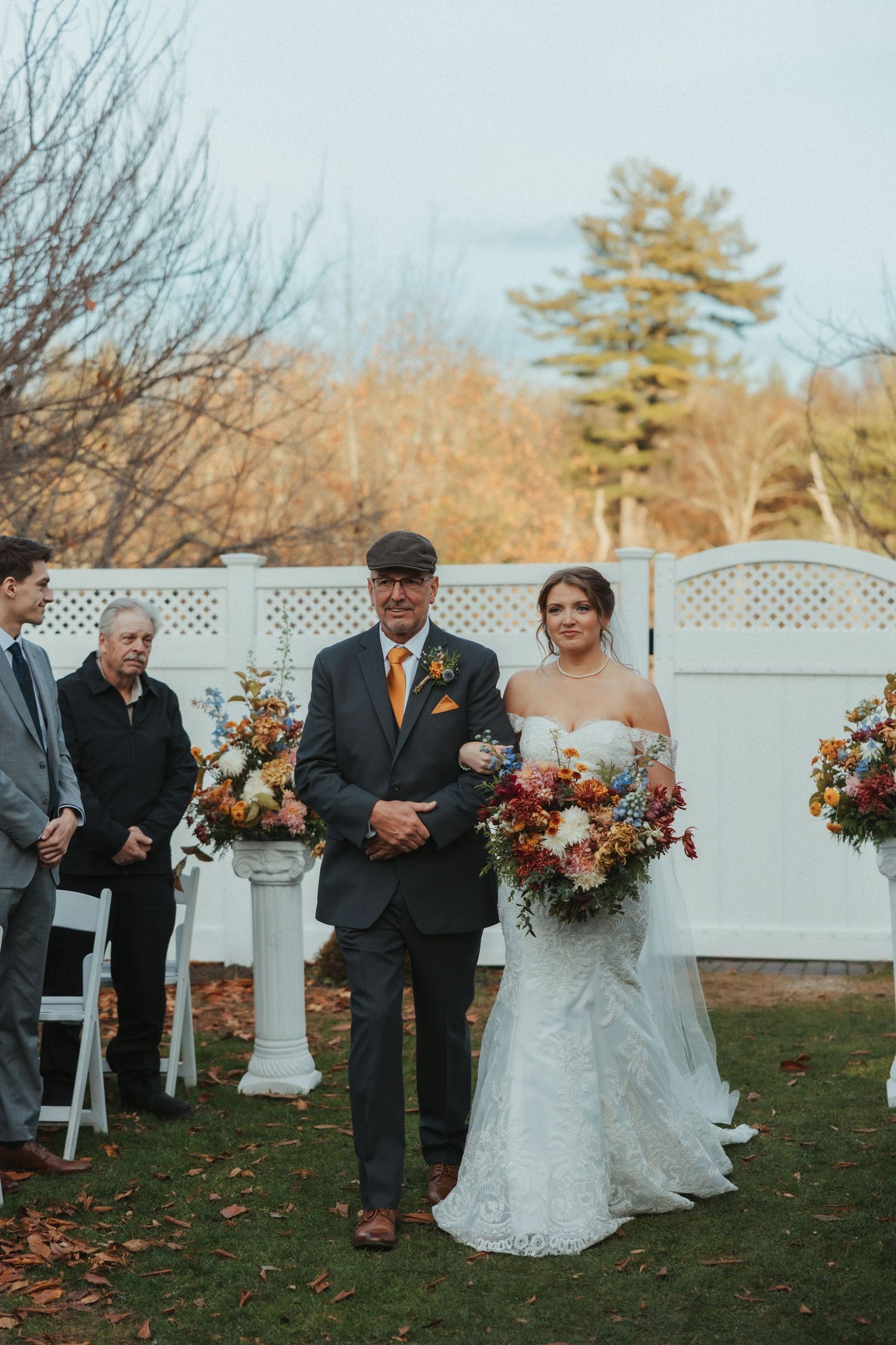 A wedding ceremony setup outdoors with white lattice fencing and autumn floral arrangements on pedestals.