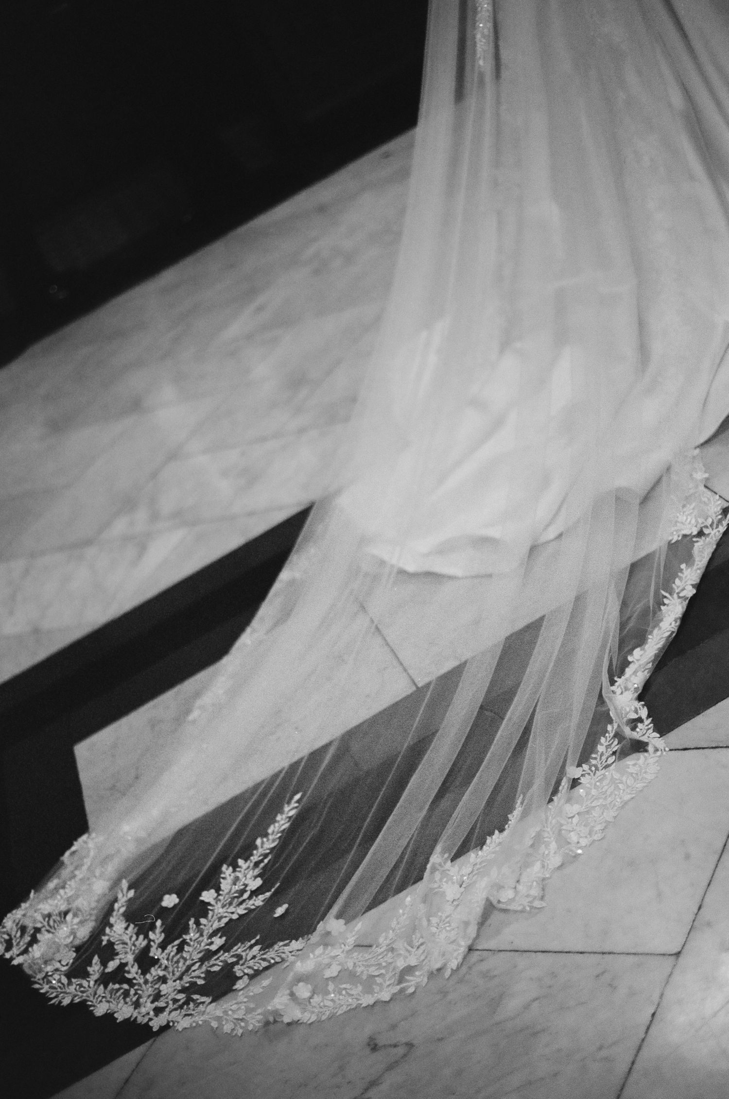 Black and white photo of an elegant bridal veil with lace trim trailing on marble floor steps.