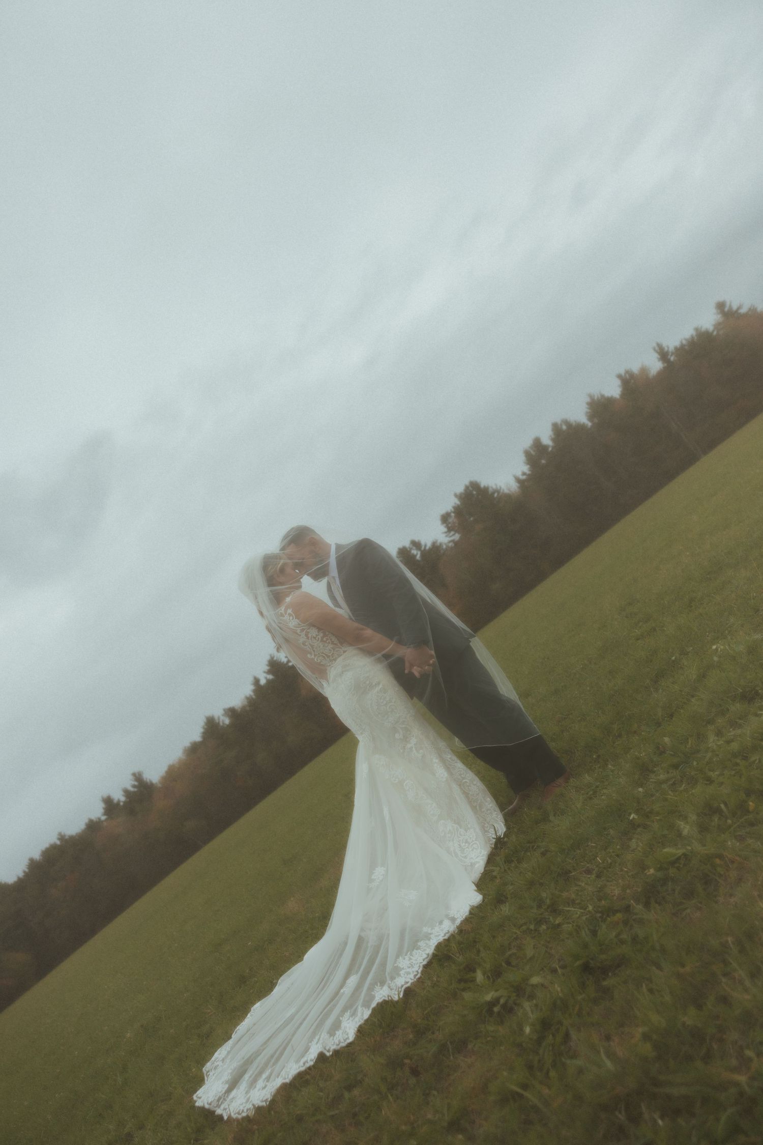 A romantic wedding couple embraces on a misty hillside with a long flowing white dress trailing down the slope.