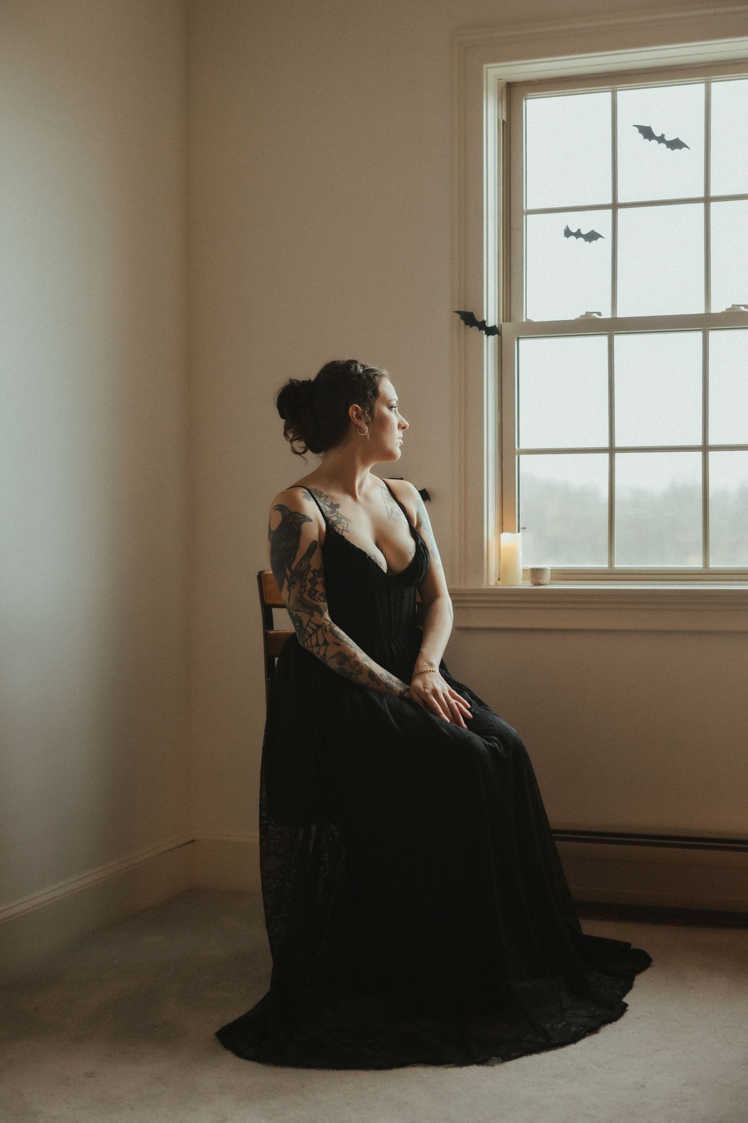 Figure in flowing black dress poses gracefully near window in minimalist room with natural lighting.