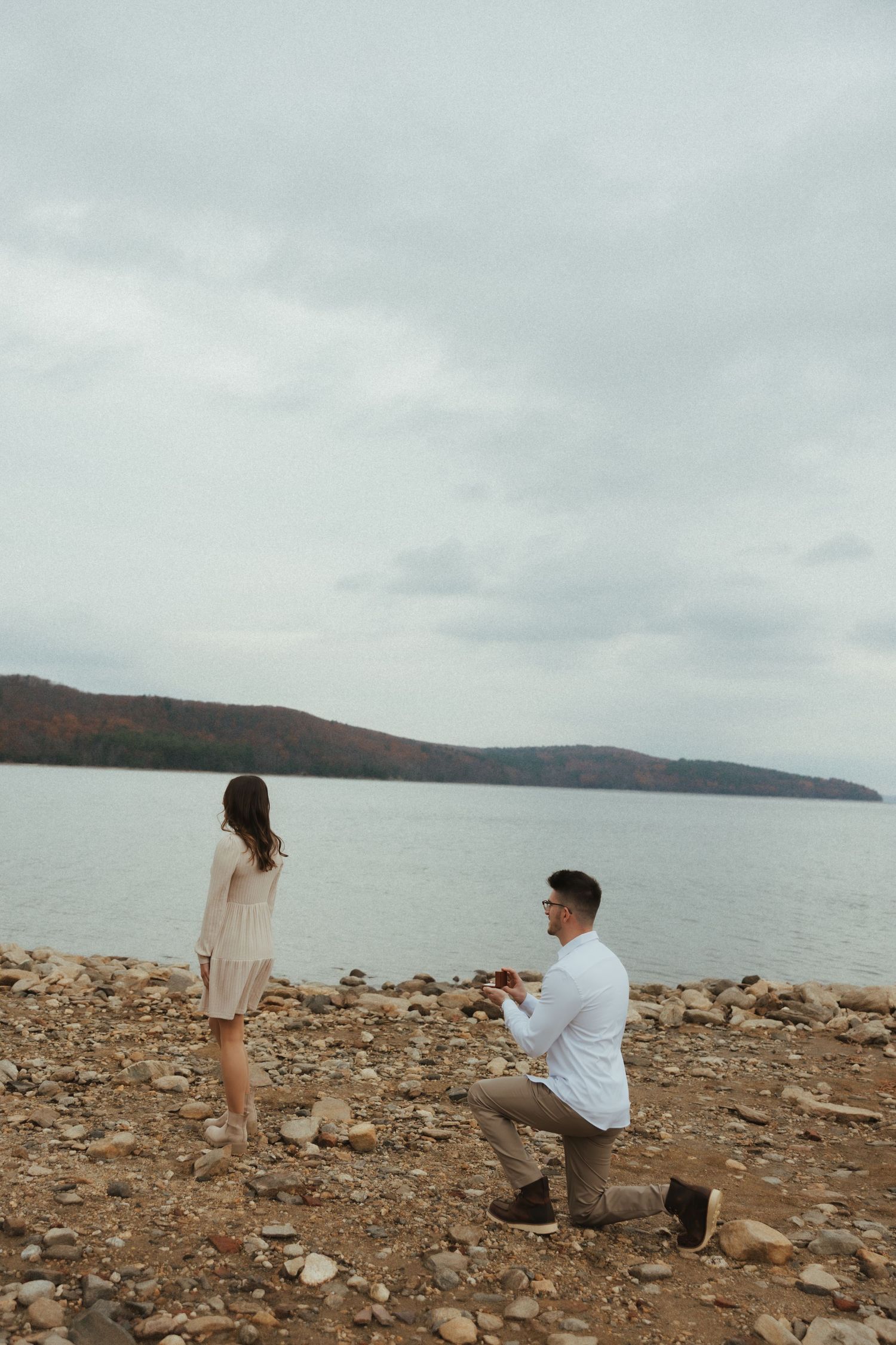 Two people share a romantic moment on a rocky beach shoreline with cloudy skies and mountains in the distance.