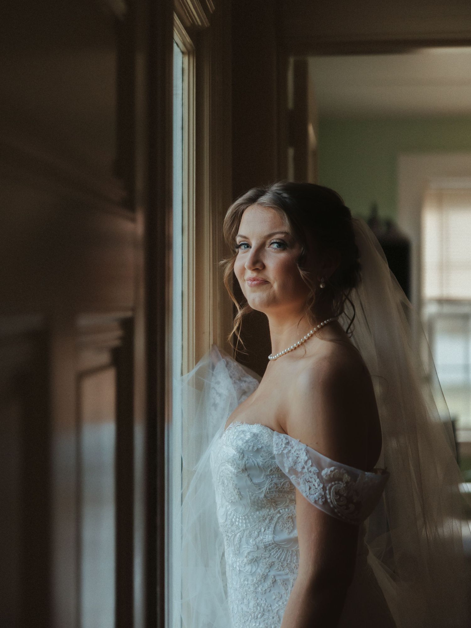 Bride in lace off-shoulder wedding gown and veil poses elegantly by window in soft natural light.