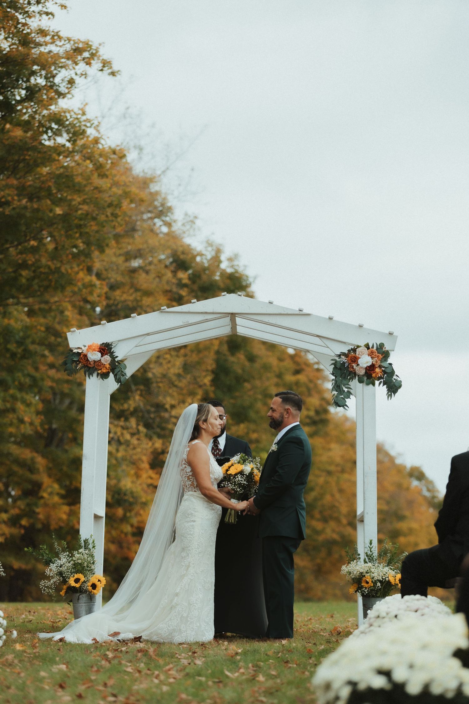 A wedding ceremony takes place under a white pergola surrounded by autumn trees with golden foliage.
