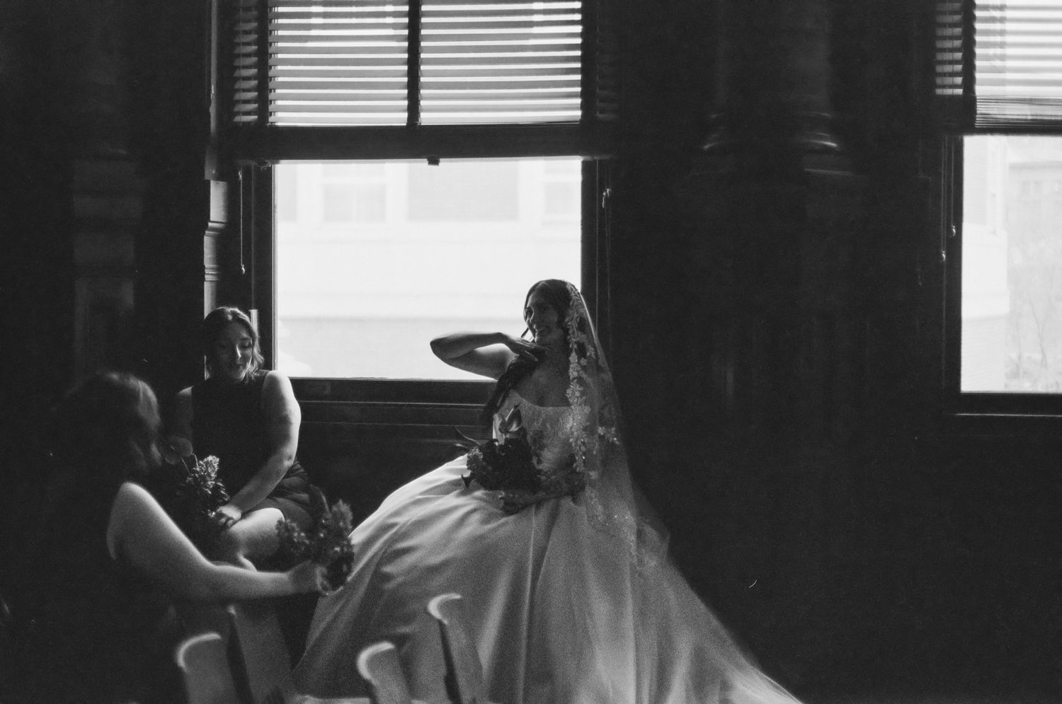 Black and white photo of a bride in a full gown sitting by large windows with bridesmaids nearby.