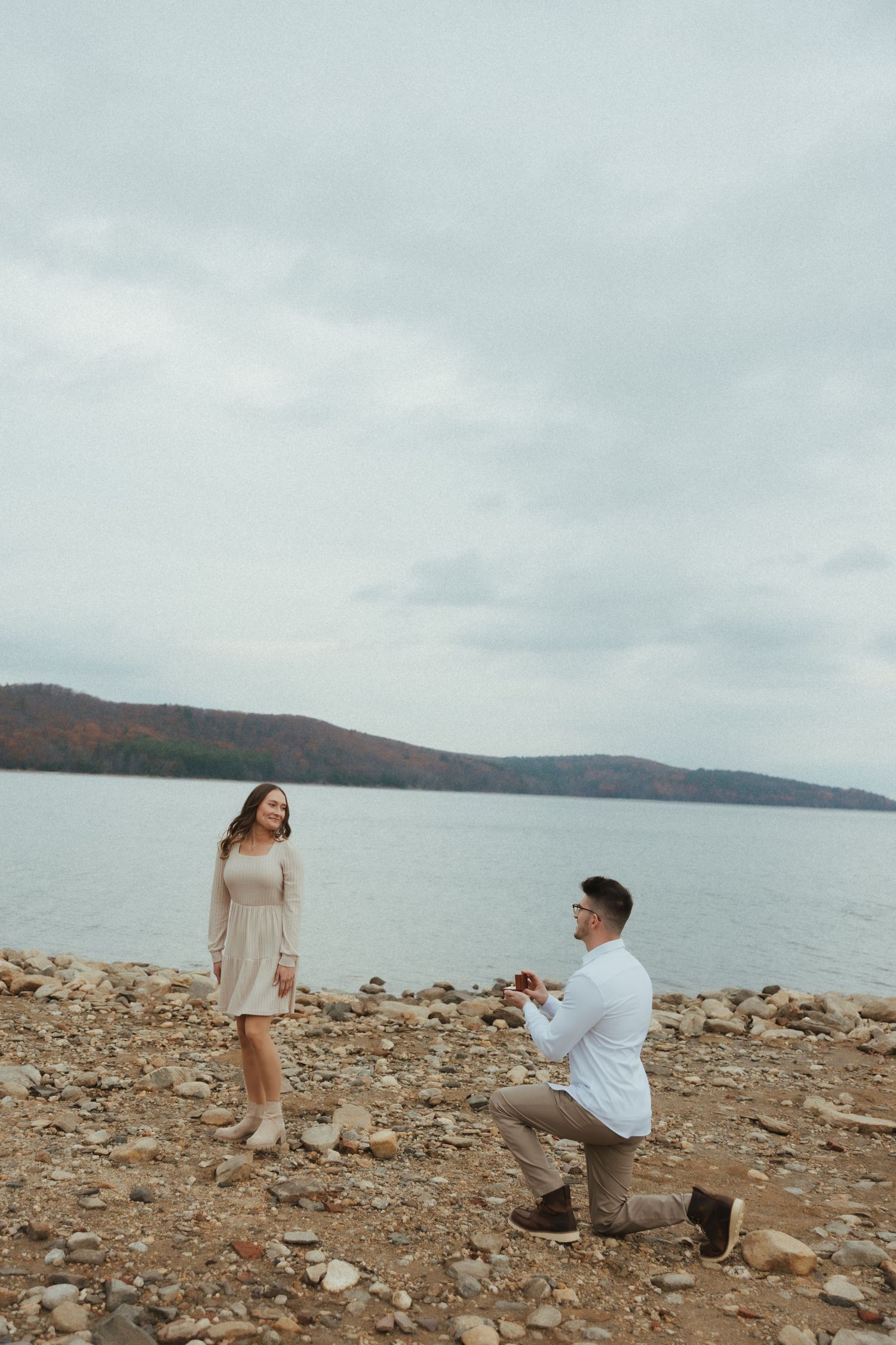 A couple sharing a romantic proposal moment on a rocky beach shore with mountains and water in the background.