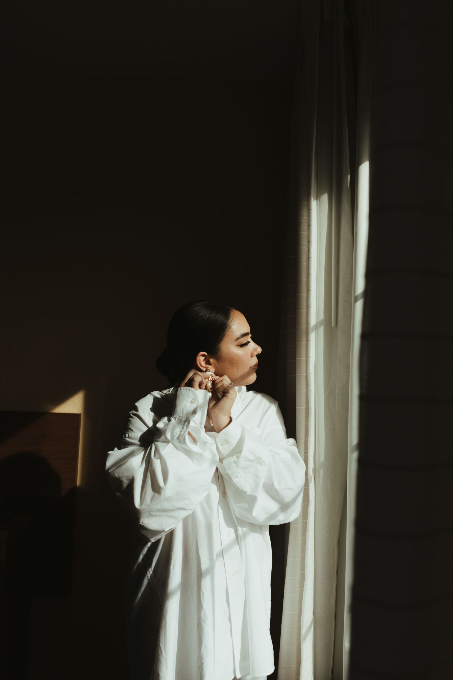 Figure in a flowing white dress stands pensively by a window with dramatic shadows and natural light.
