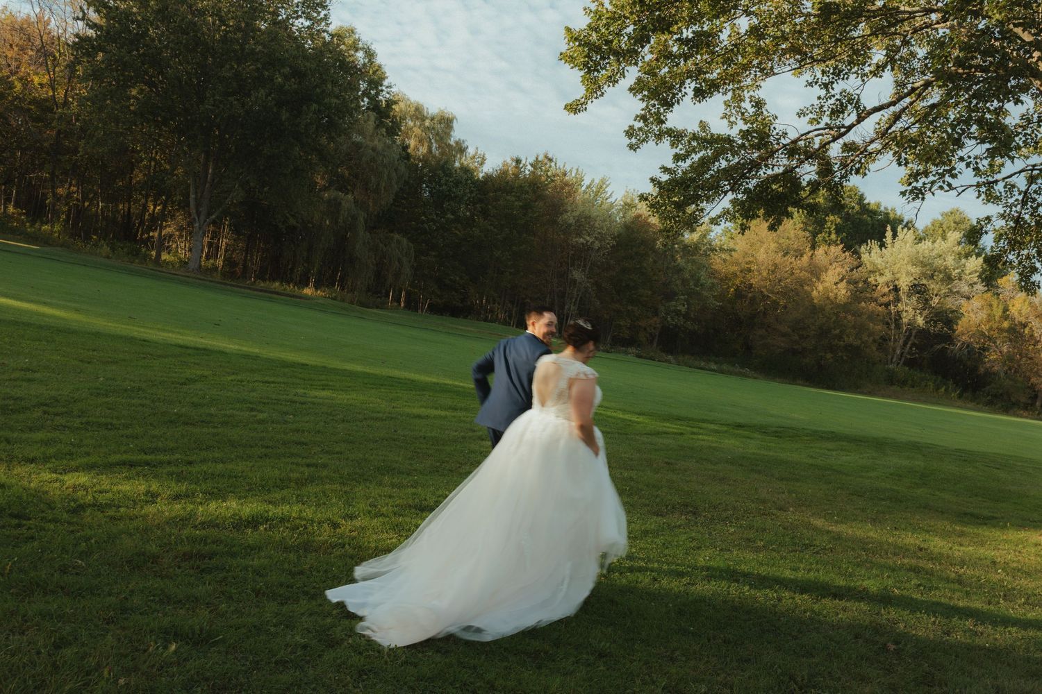 A couple in formal wedding attire walk across a sunlit green lawn with autumn trees in the background.