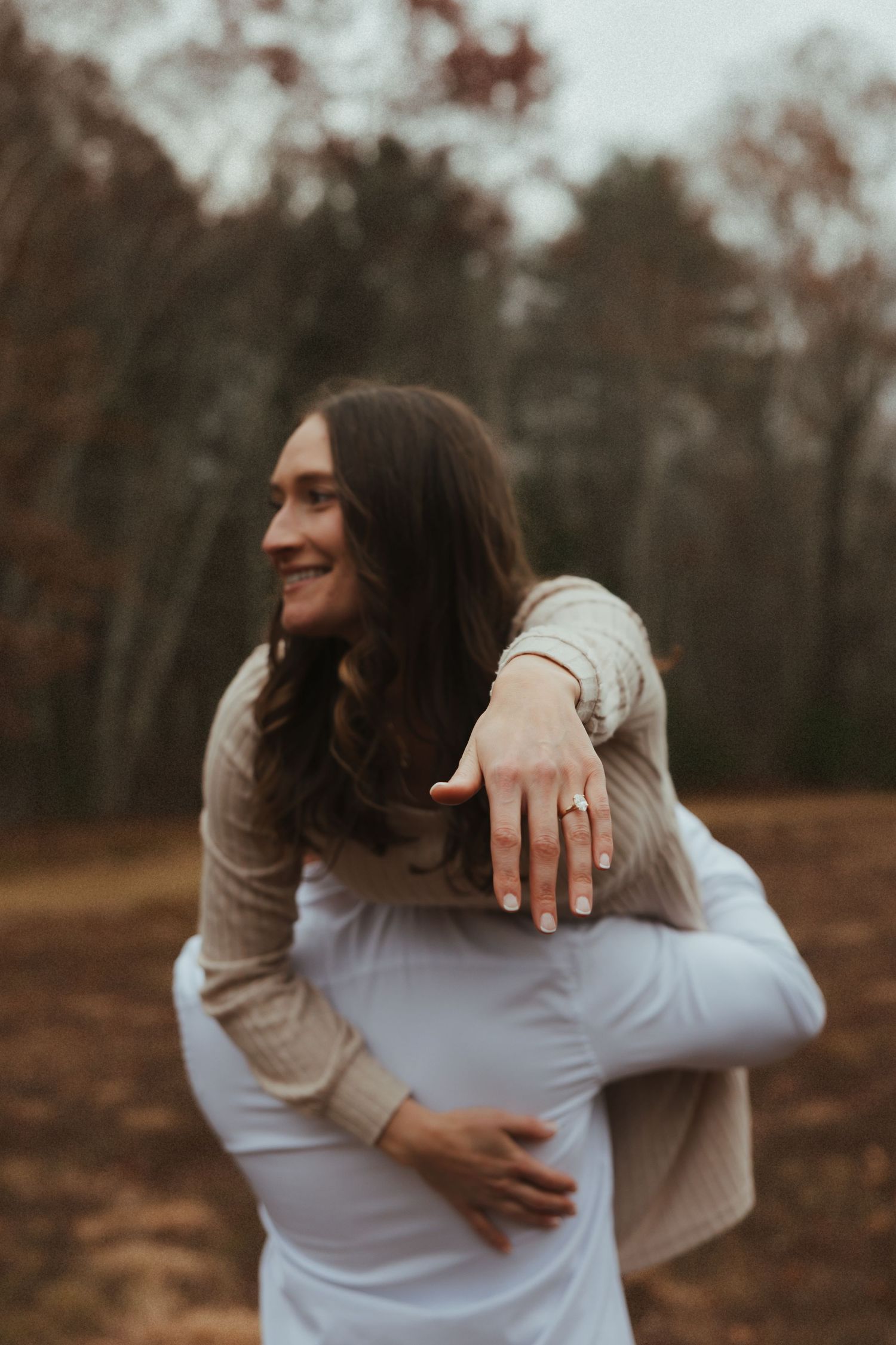 Two people in white and beige clothing embrace in a tender moment against an autumn forest backdrop.