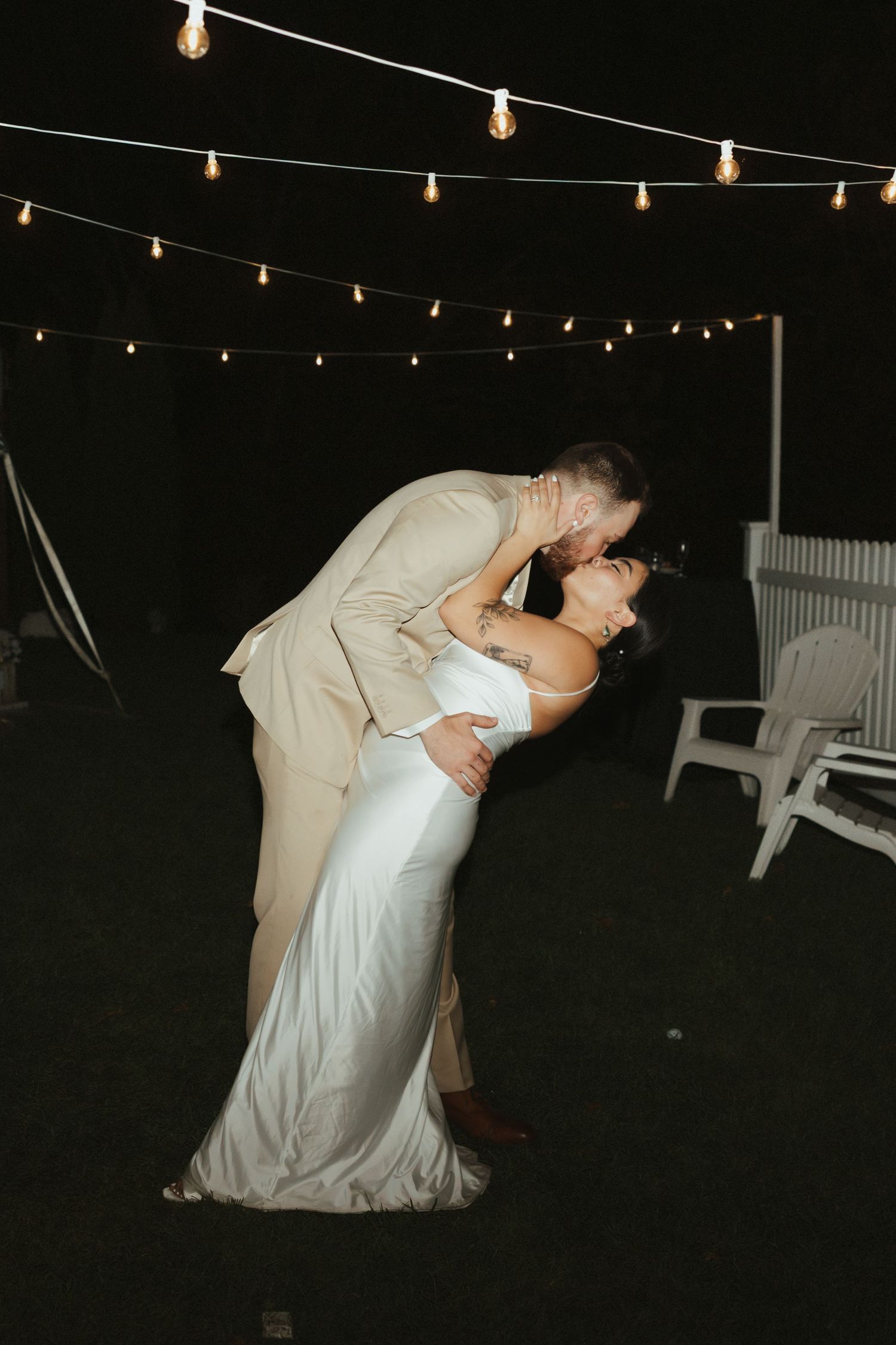A couple in wedding attire share a romantic dip under twinkling string lights during their evening celebration.