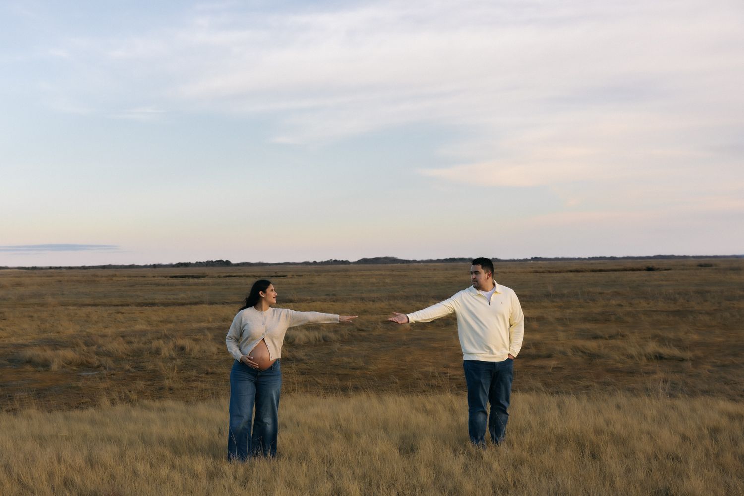 Pregnant couple holding hands in a golden grass field at dusk under a pastel sky.