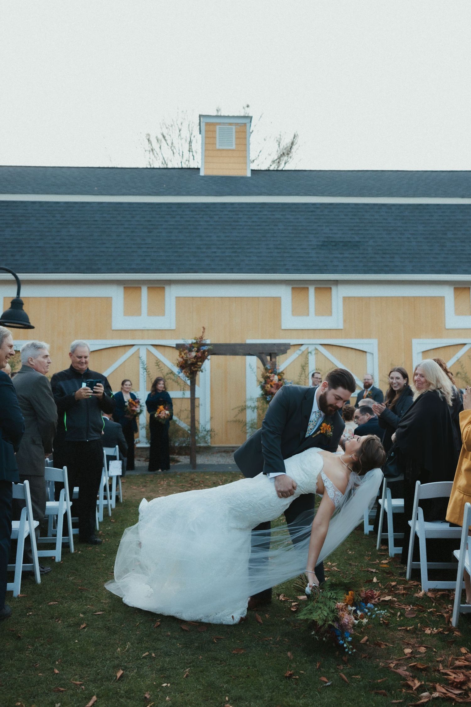 A couple shares a romantic dip kiss at their outdoor wedding ceremony with guests watching and white chairs arranged on the lawn.
