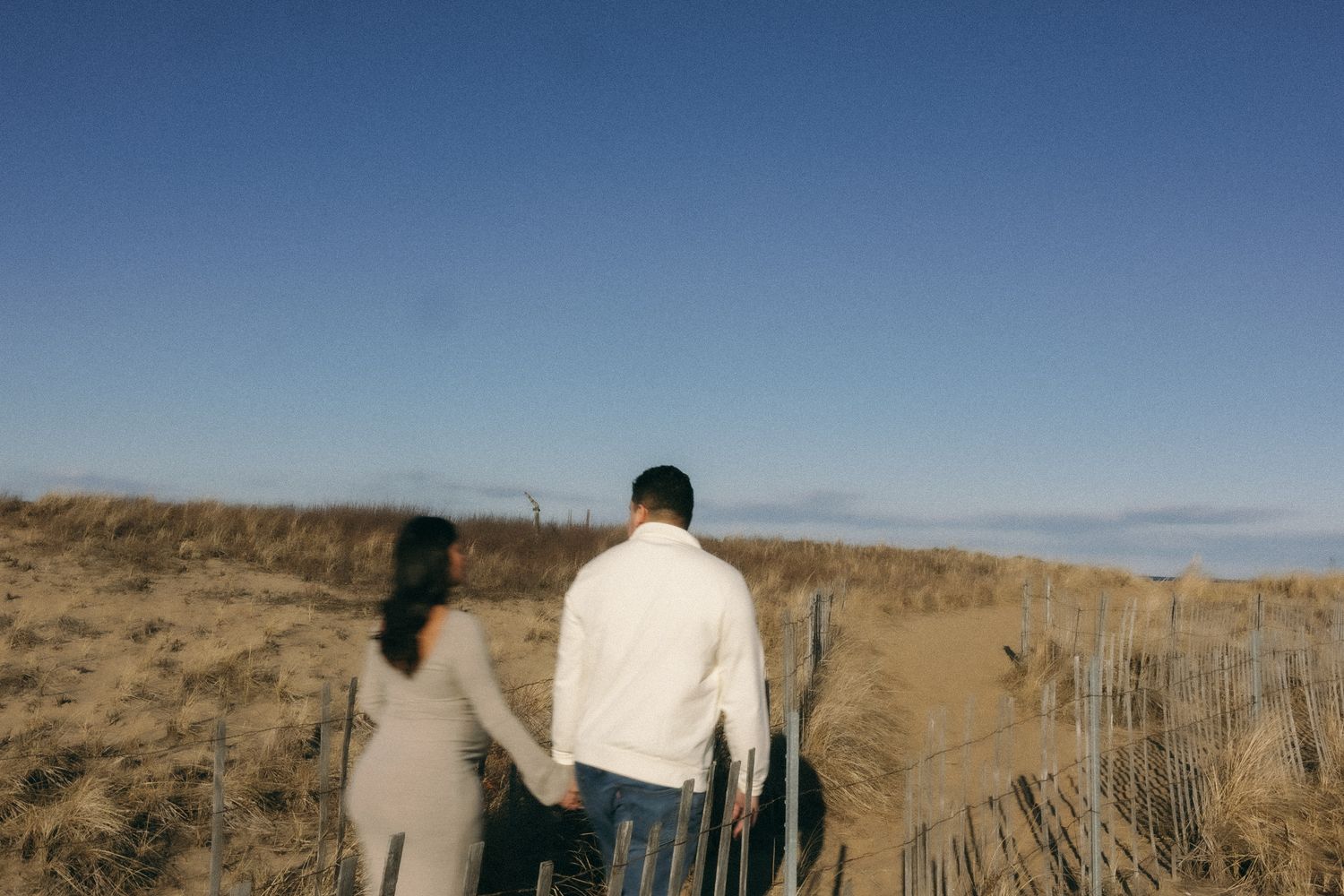 Couple holding hands walking along a sandy beach path with wooden fence under a clear blue sky.