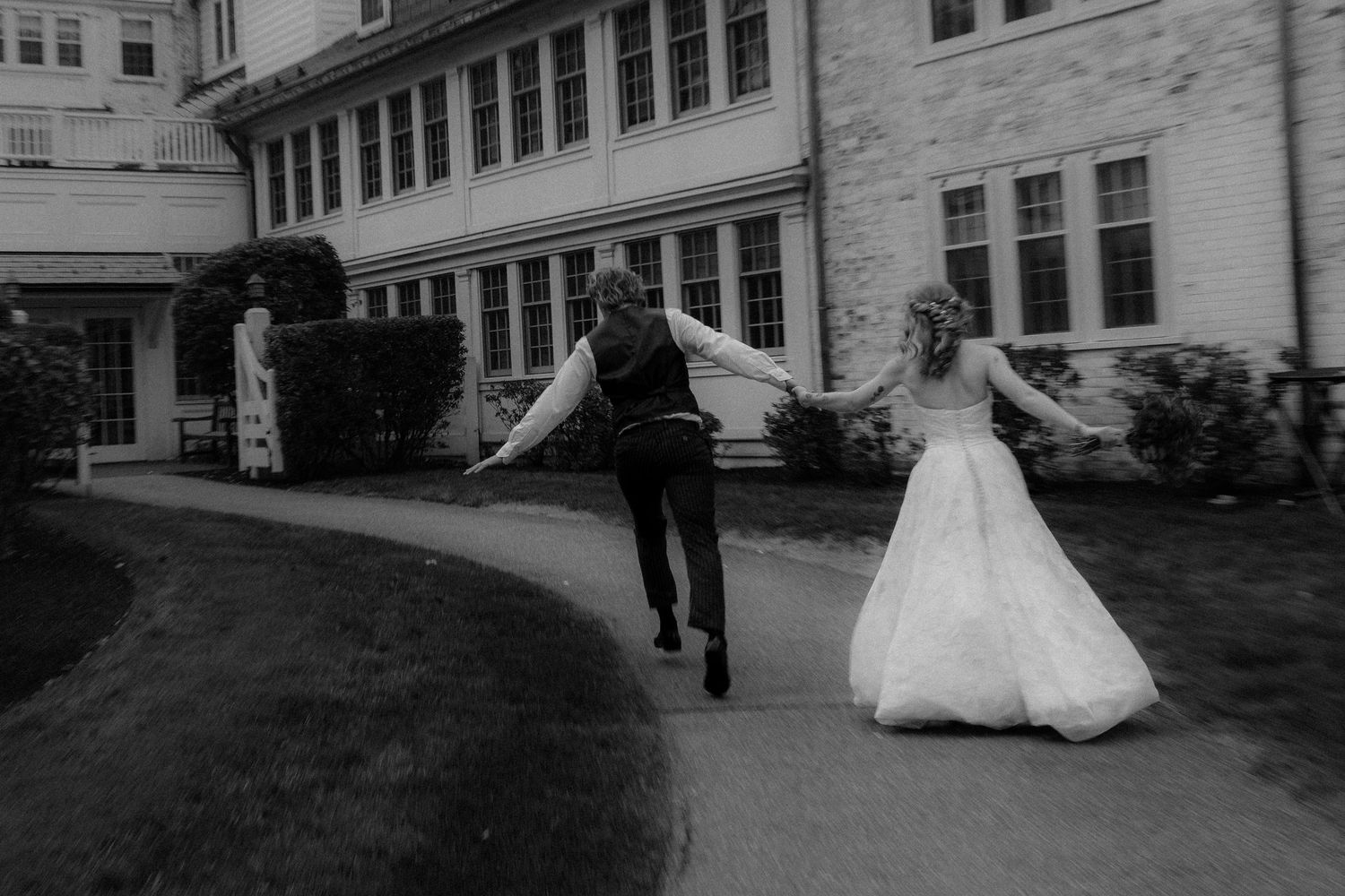 A joyful couple in wedding attire runs together down a curved path in front of a historic white building.