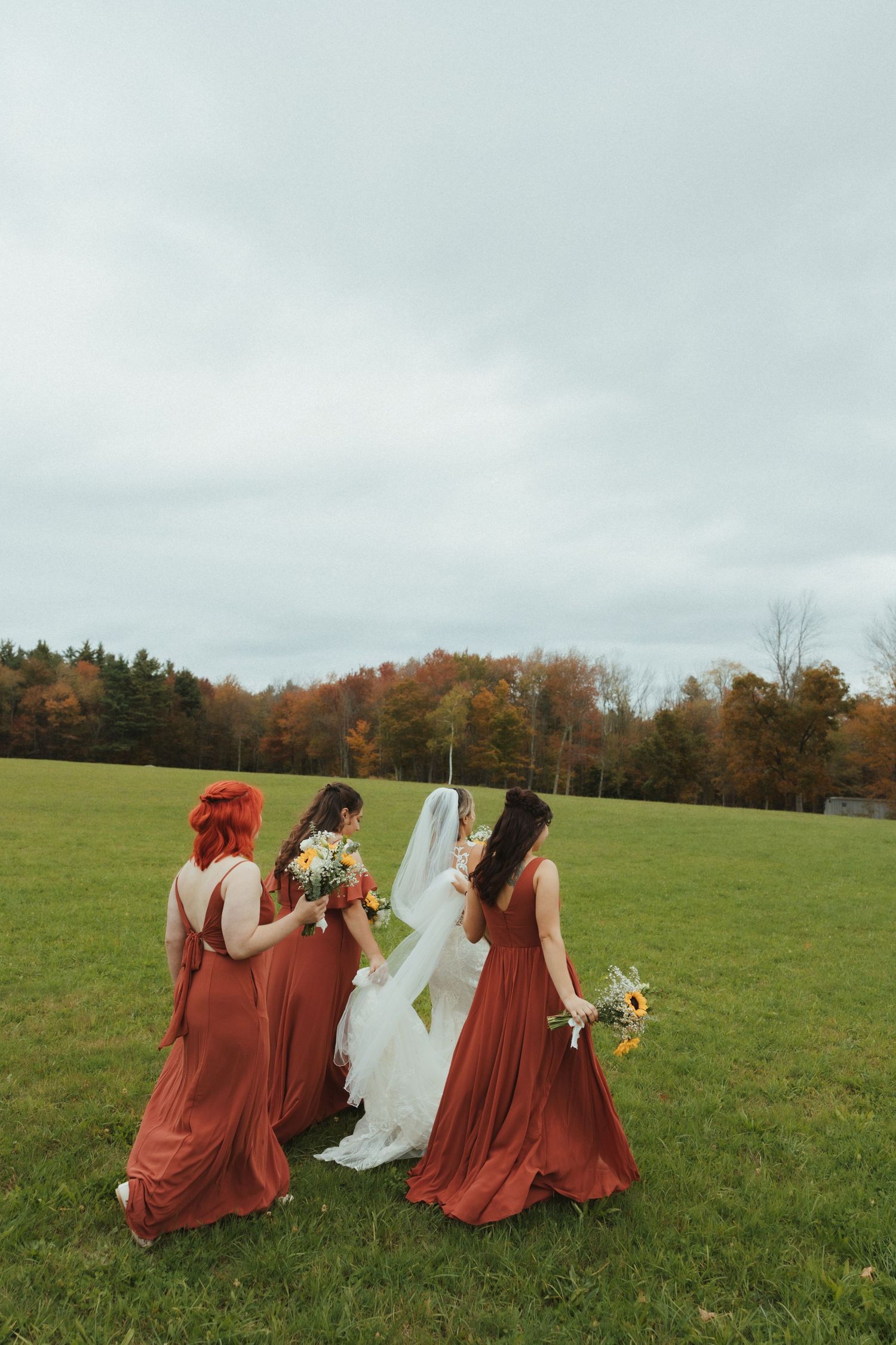 Bridesmaids in rust-colored dresses walk with a bride across a green field on an overcast autumn day.