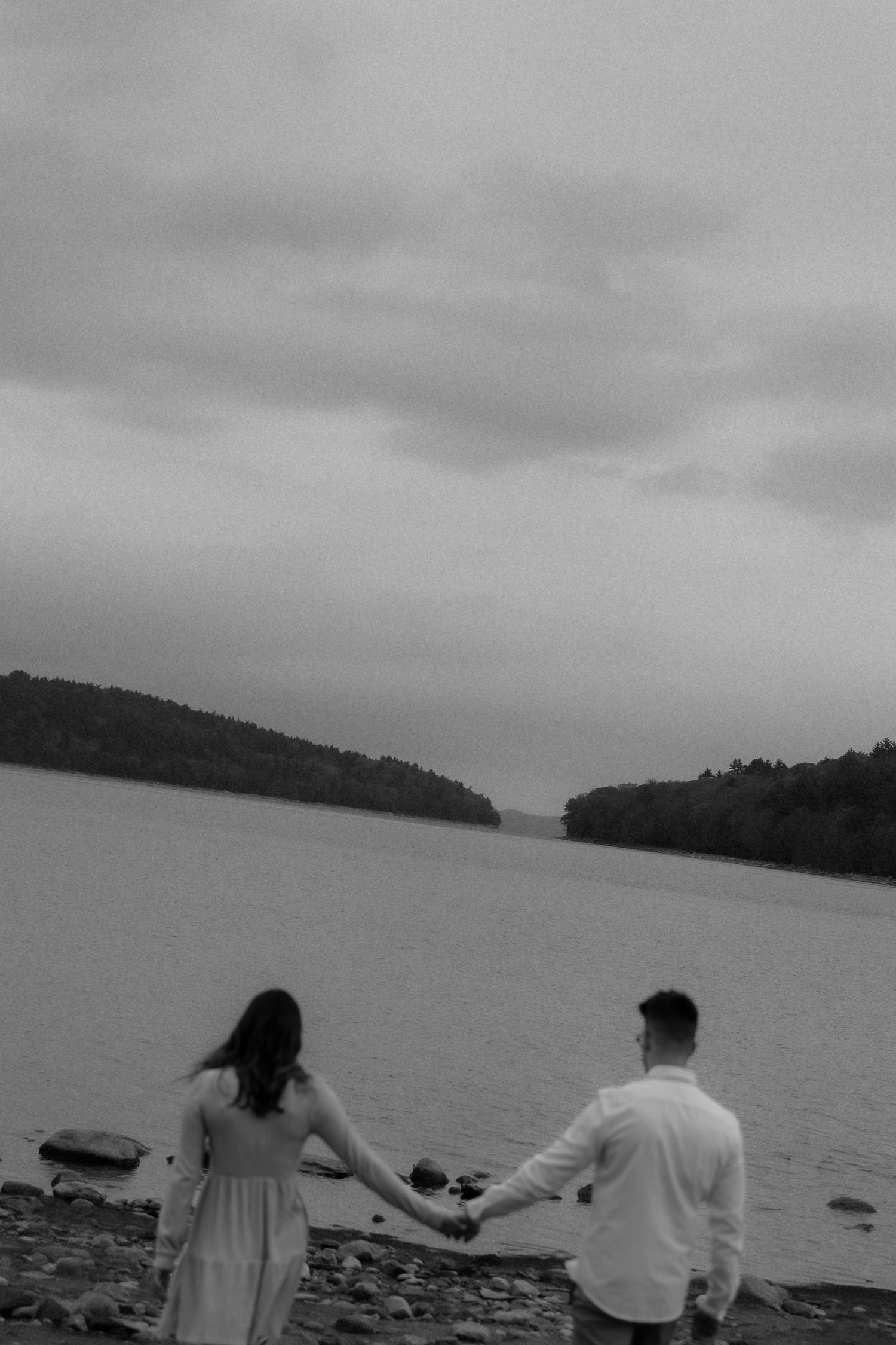 Two people hold hands while walking along a rocky shoreline with a misty lake and forested islands in the background.
