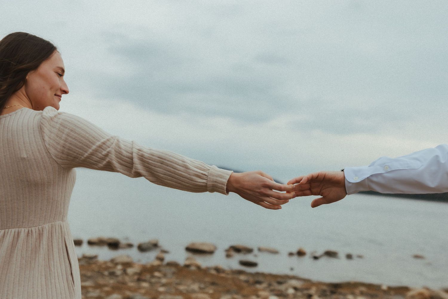 Two hands reaching out to connect on a rocky beach with mountains and cloudy skies in the background.