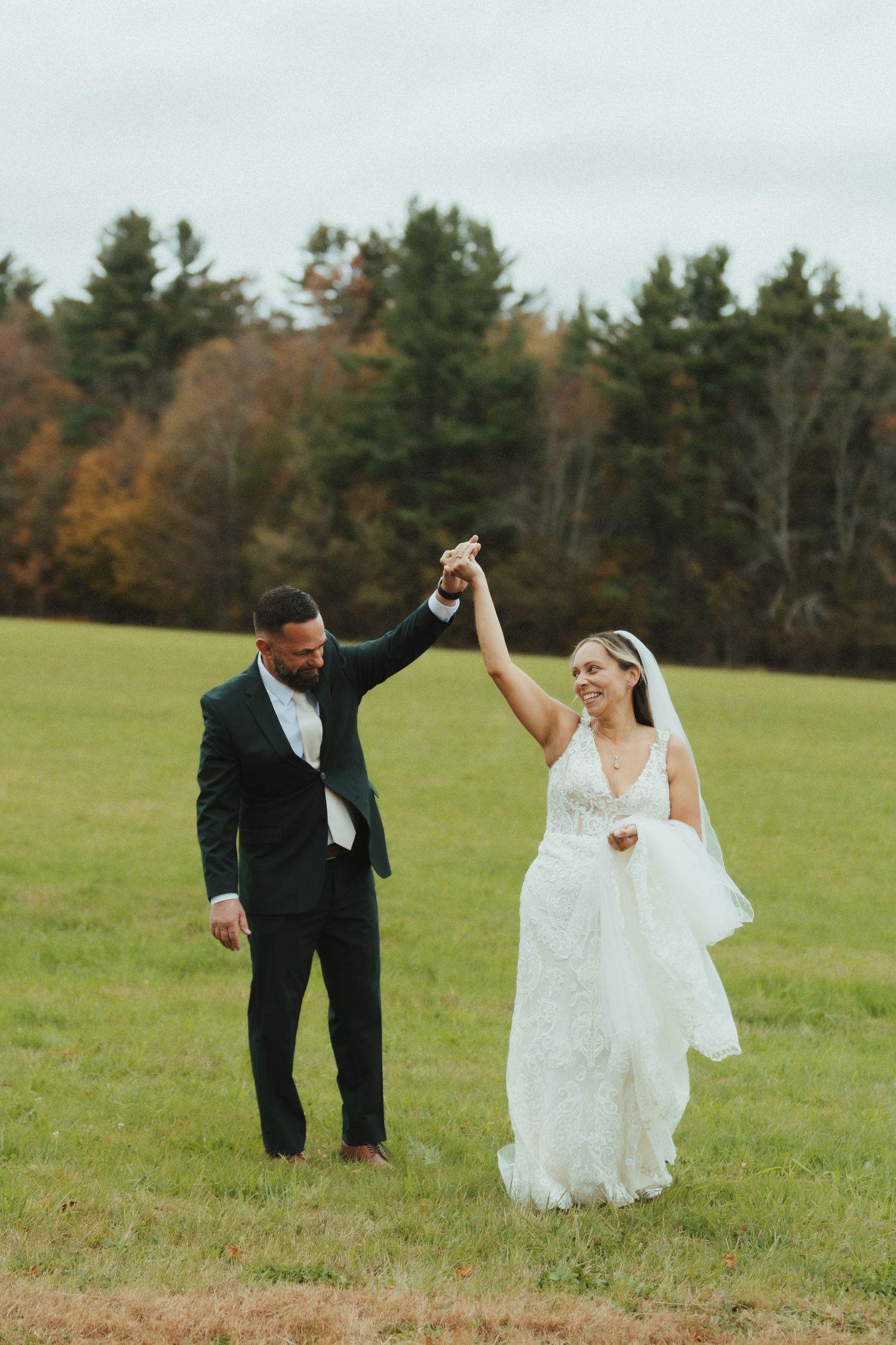 A couple in wedding attire dances and twirls together on a grassy hillside with autumn trees in the background.