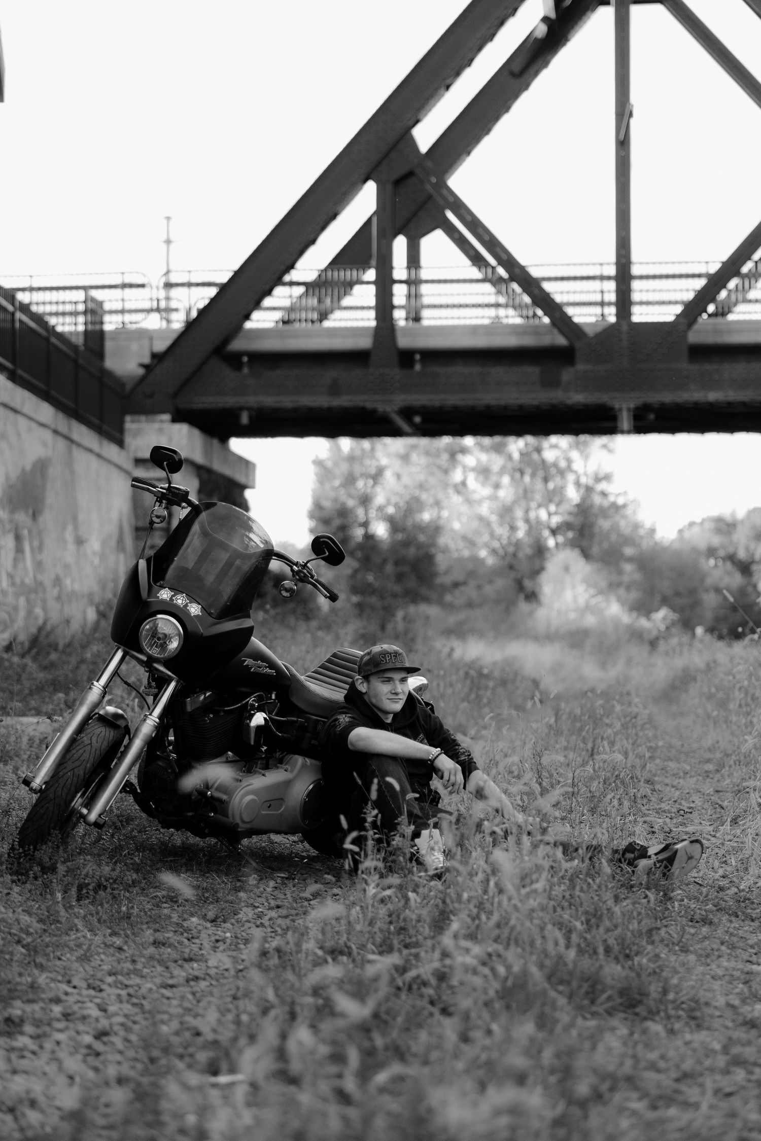 Black and white photo series showing a motorcycle laid down in tall grass near a railroad bridge structure.