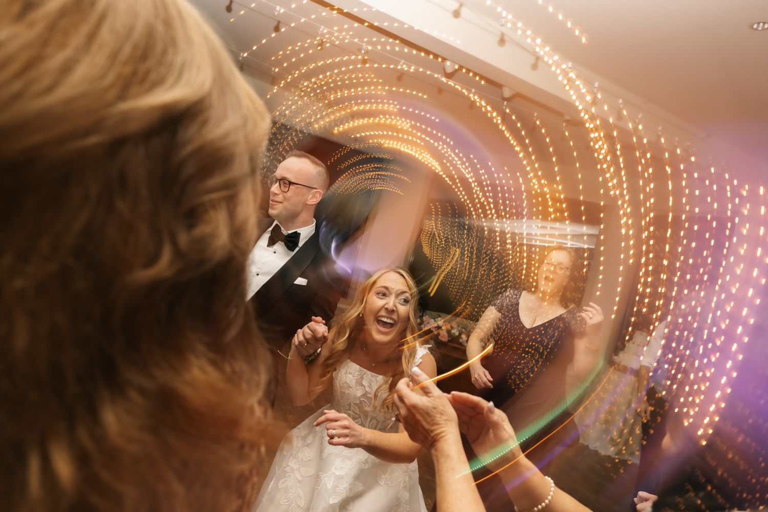 Bride laughing joyfully on wedding dance floor surrounded by guests, with swirling bokeh lights creating magical atmosphere.