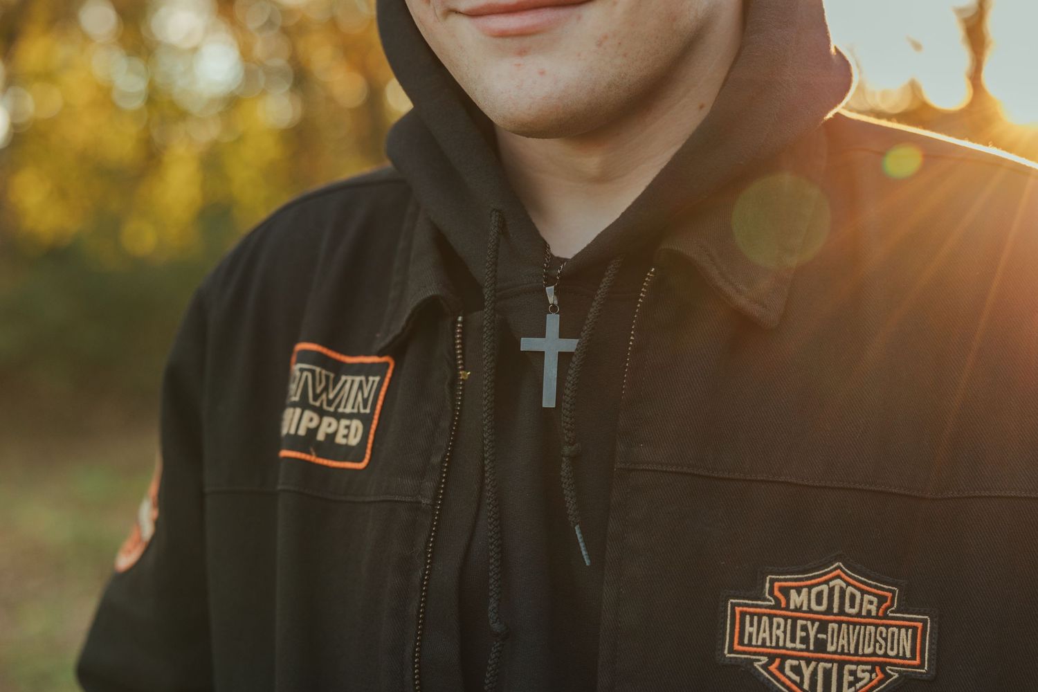 Person wearing black Harley Davidson jacket with cross necklace at sunset with golden bokeh background.