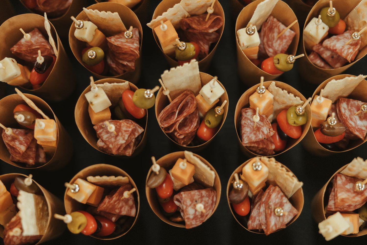 Overhead view of individual appetizer cups filled with cured meats, cheese cubes, and olives arranged in rows.