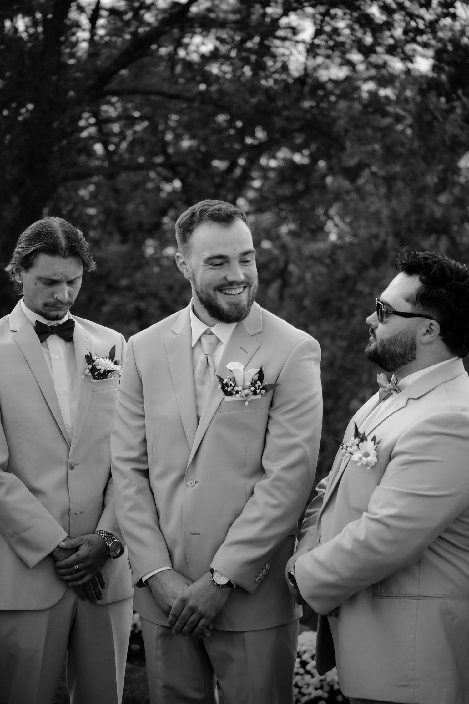 Groomsmen in light beige suits stand together in a candid moment sharing laughs at an outdoor wedding.