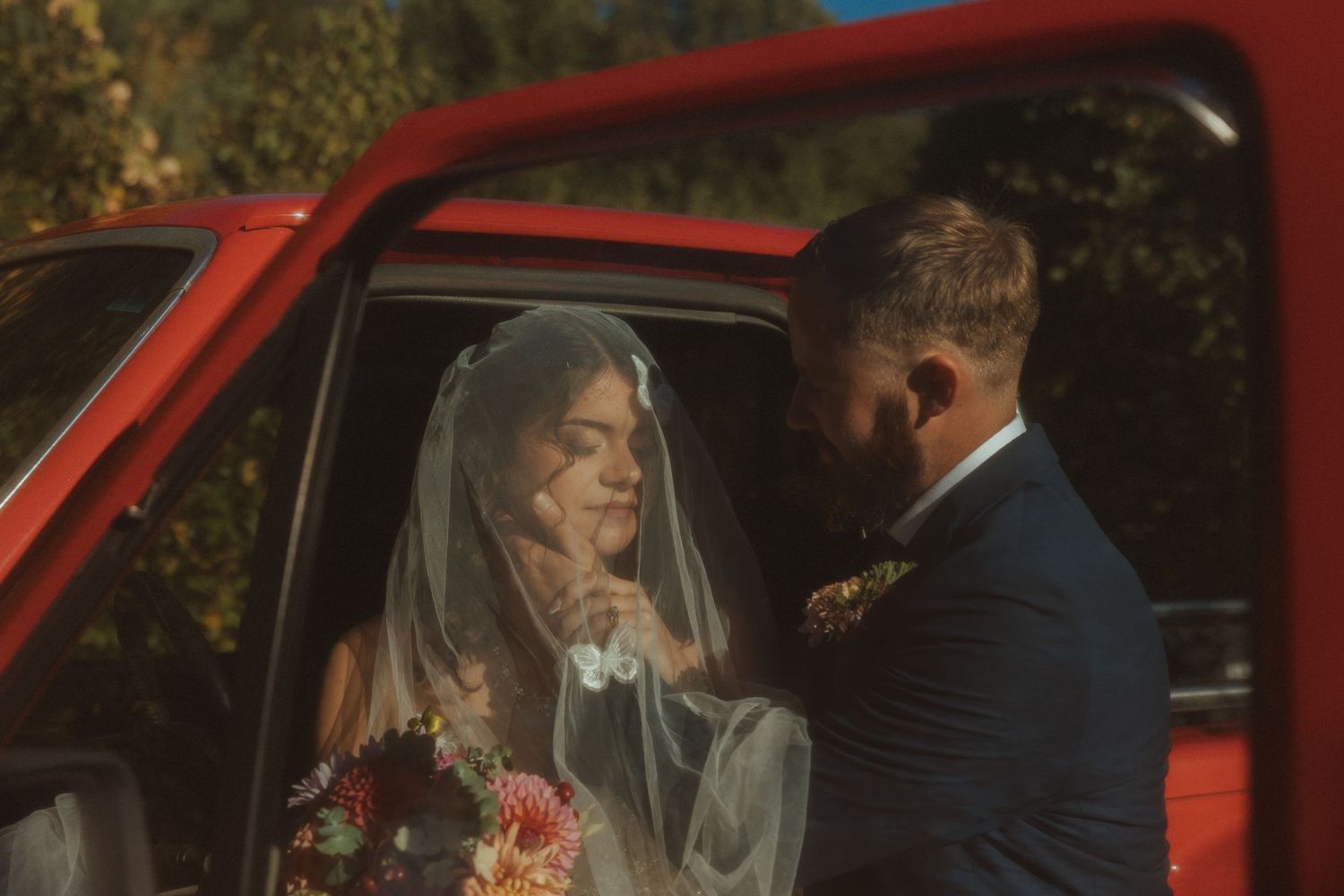 A couple sits in a vintage red car during what appears to be a wedding celebration.