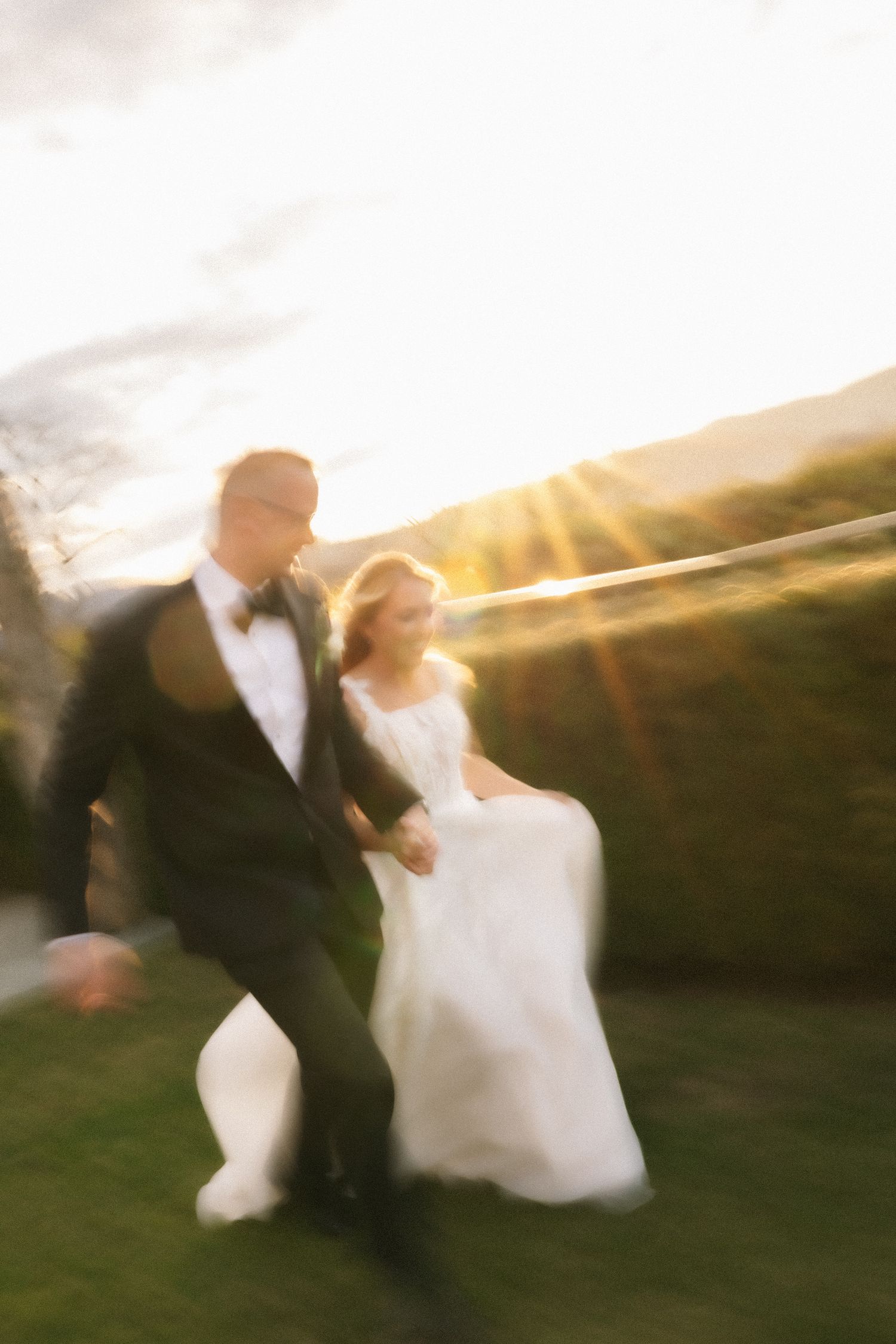 Bride and groom running joyfully on green lawn at golden sunset, motion blur capturing romantic wedding day movement.