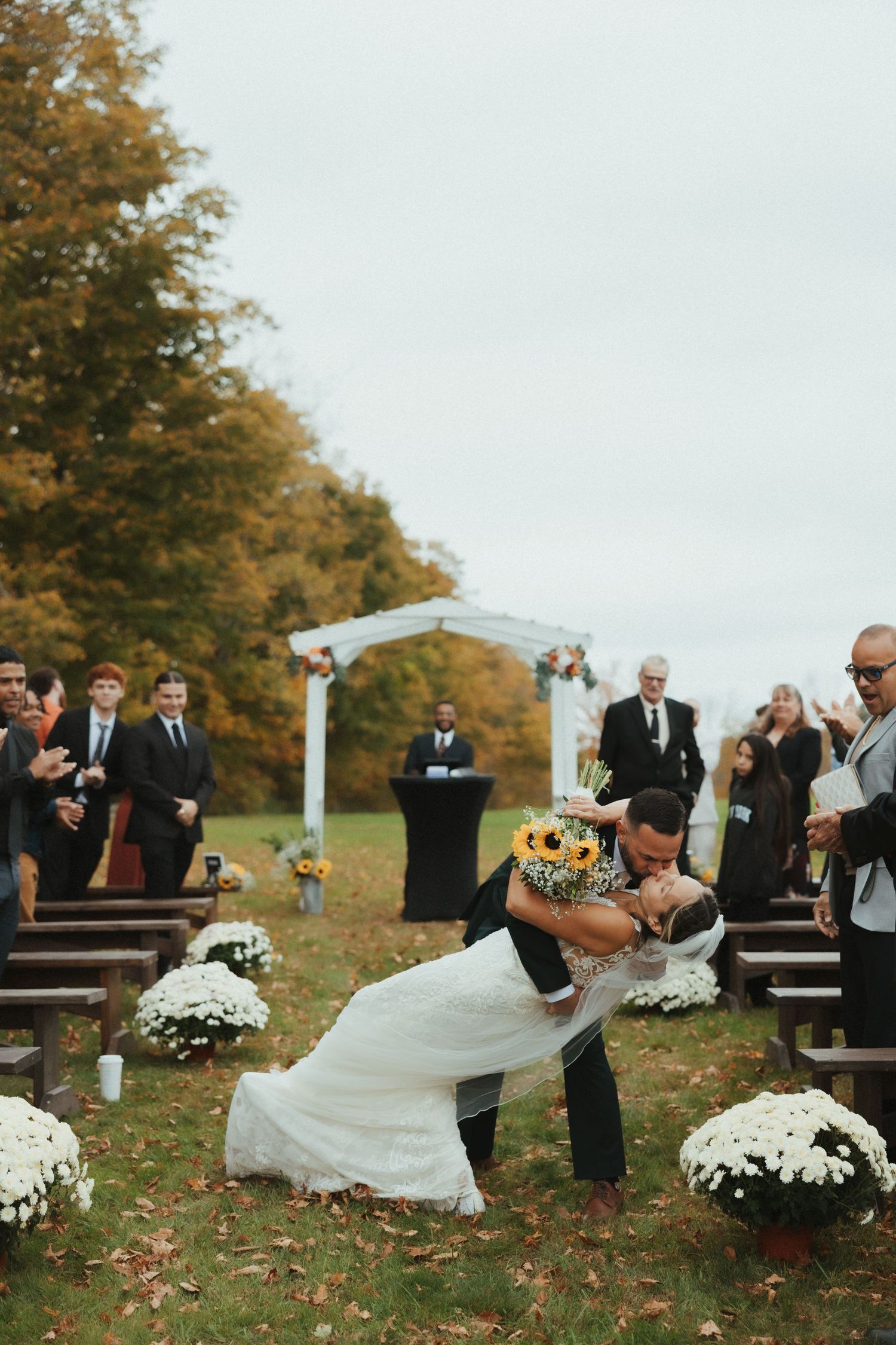Wedding guests react with surprise and laughter as strong wind blows through an outdoor autumn ceremony.