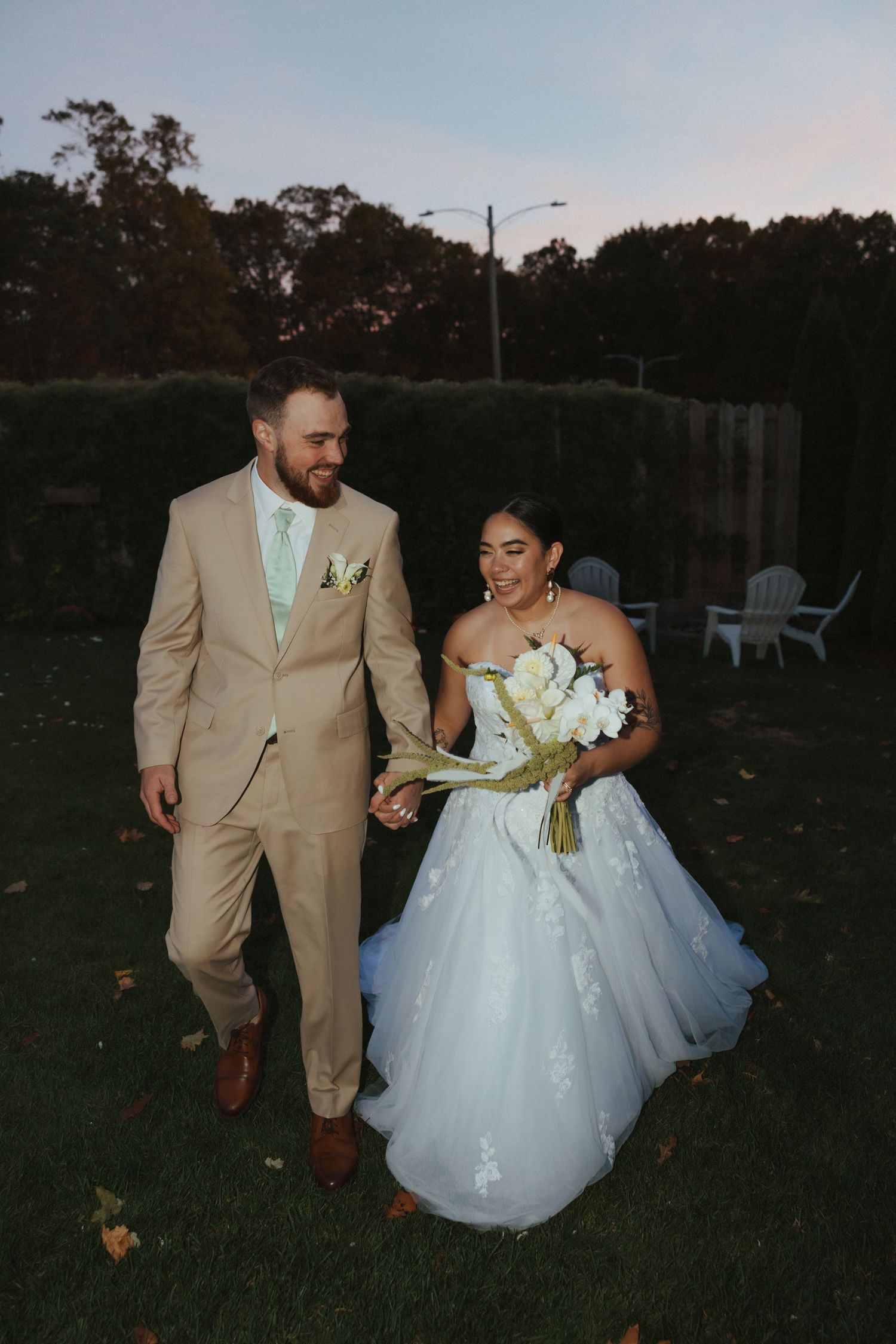 A couple in formal wedding attire walks together on grass during sunset, with the bride in white and groom in tan.