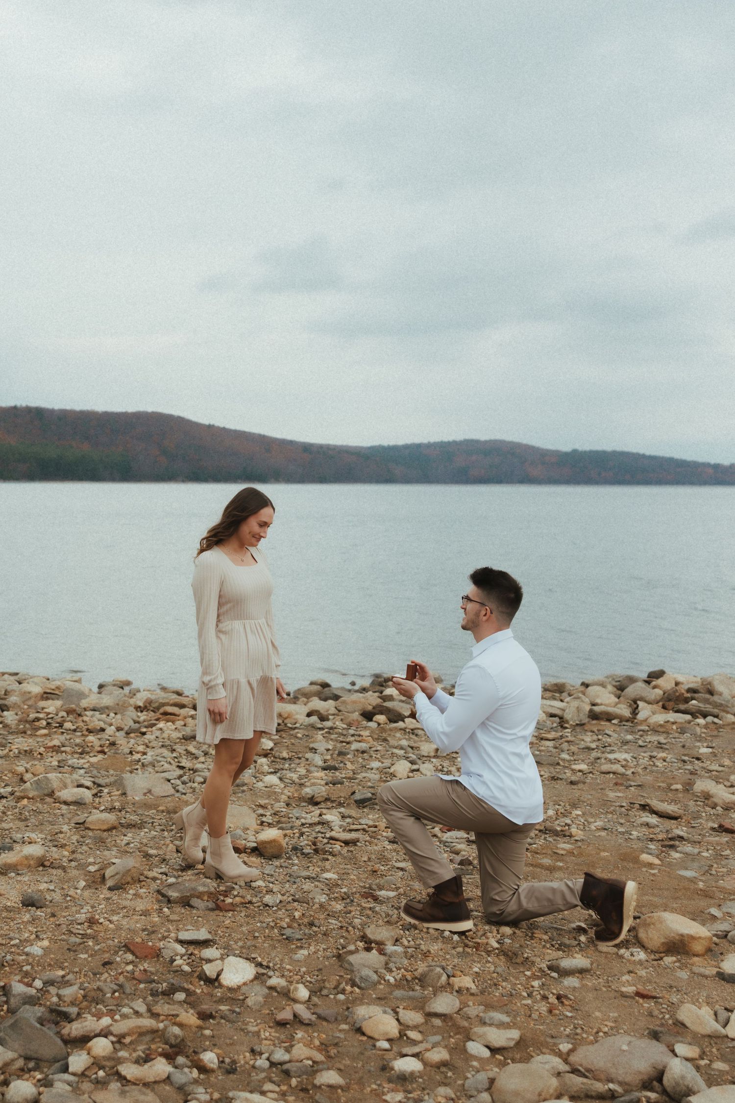 A romantic marriage proposal scene on a rocky beach shore with mountains and water in the background.