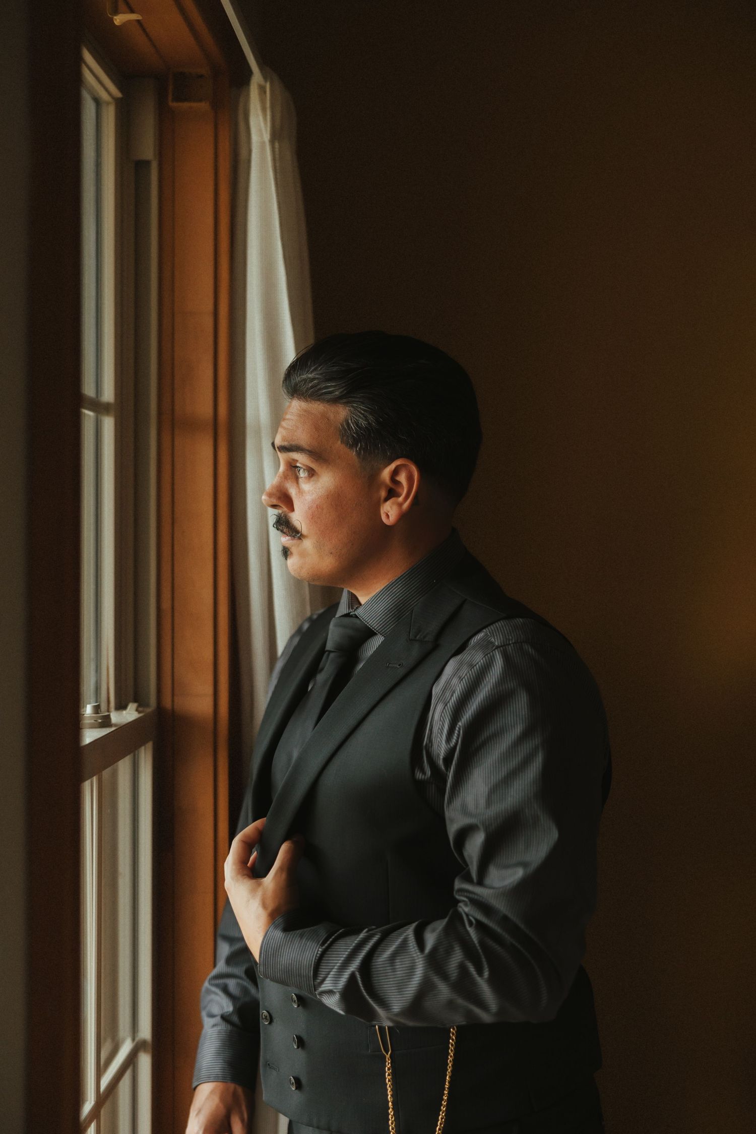 A person in a dark suit gazes contemplatively through a window in warm natural lighting.