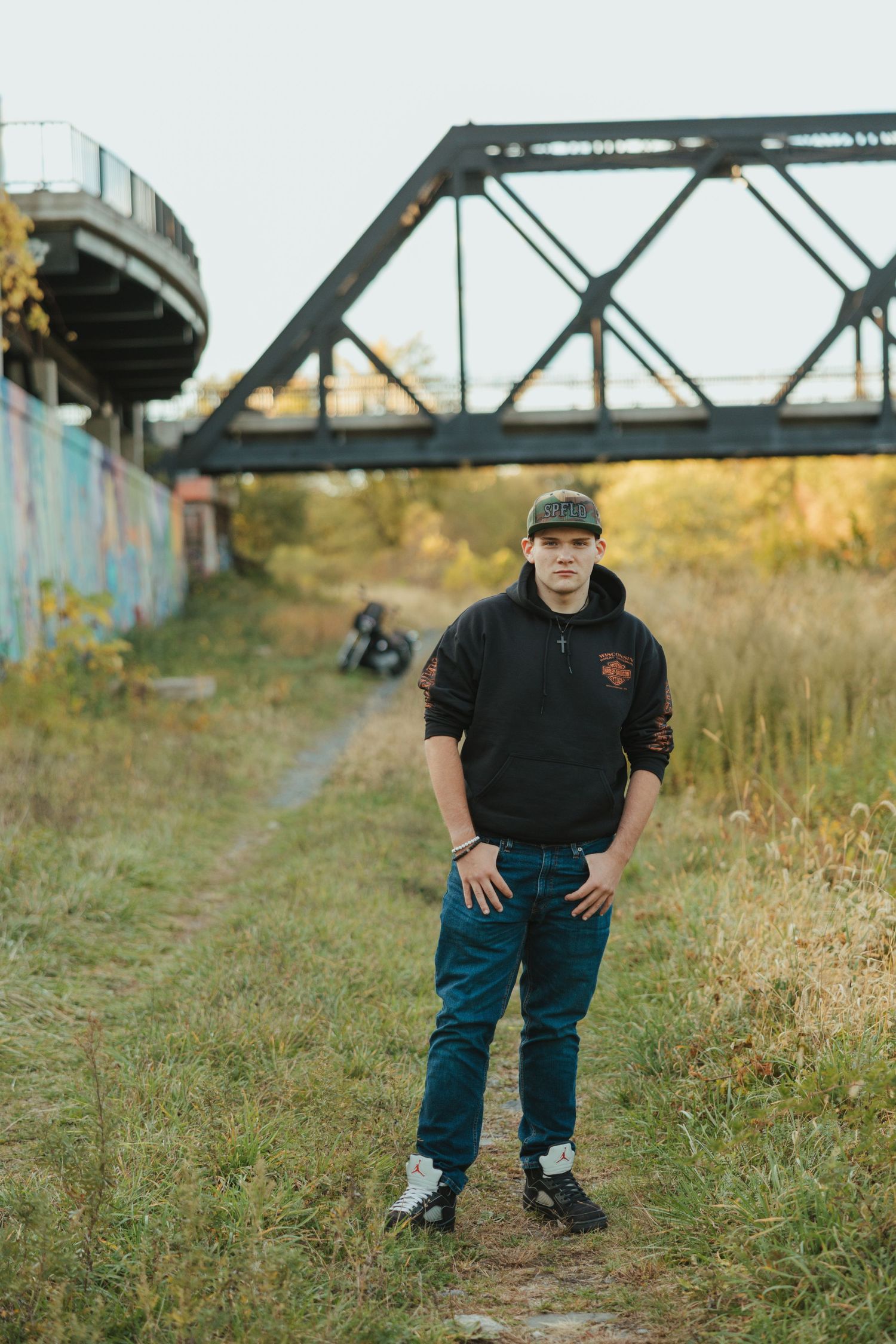 Person in dark clothing poses near a railroad bridge structure in a grassy outdoor setting.
