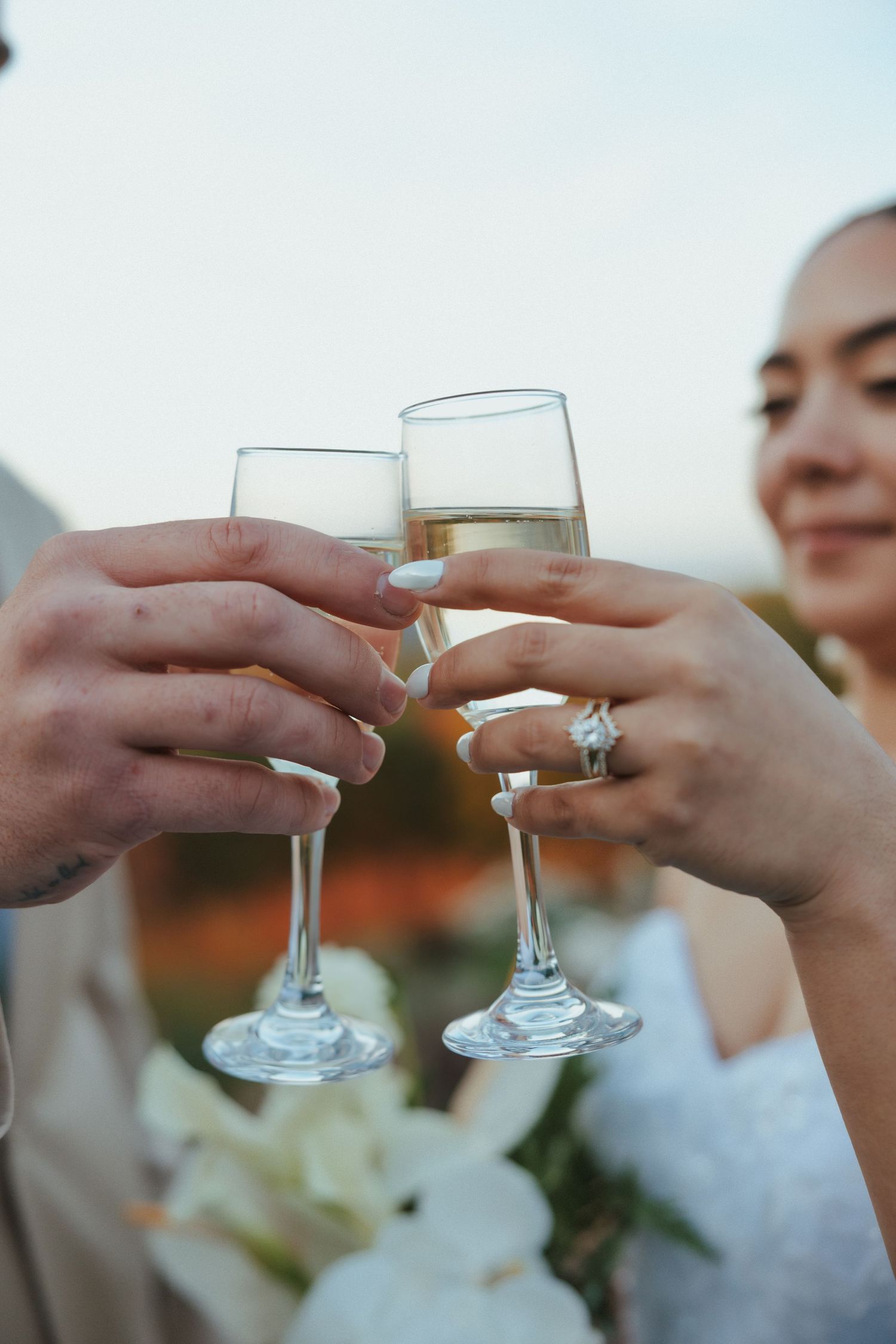 Wedding guests clink champagne glasses together in a celebratory toast against a bright sky.