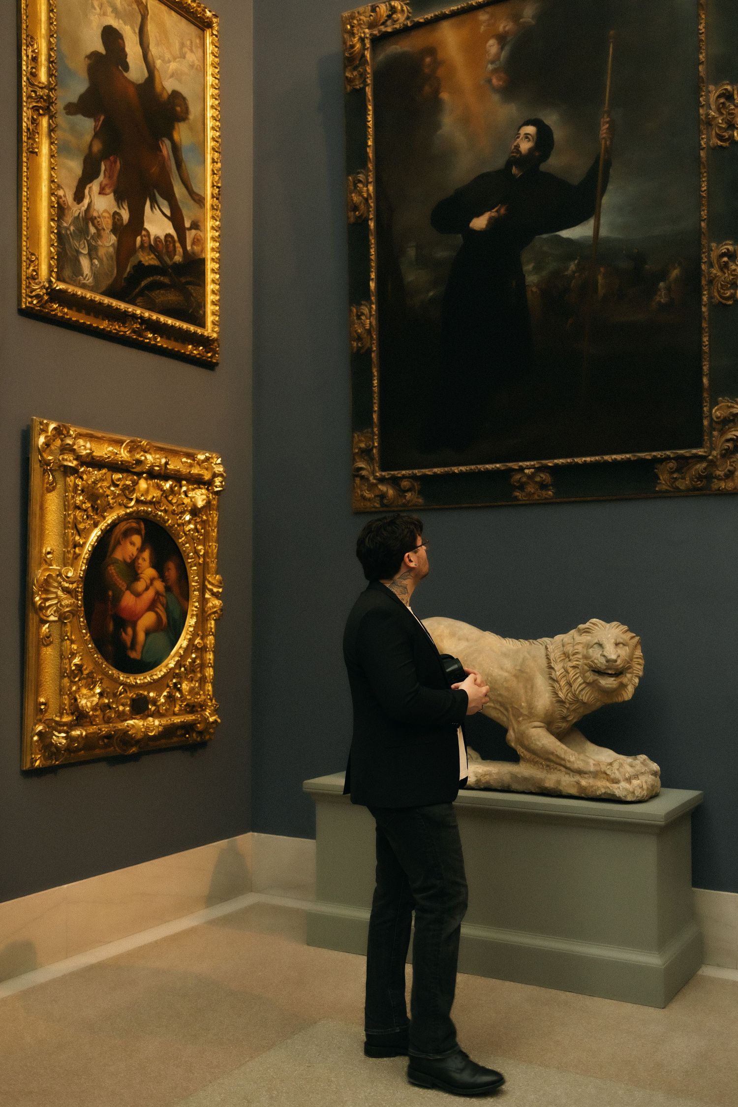 Person in black suit viewing classical paintings and a lion sculpture in an elegant art museum gallery.