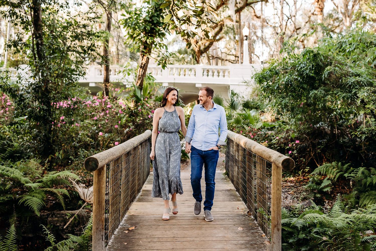A couple walks hand-in-hand across a rustic wooden bridge surrounded by lush greenery and trees.