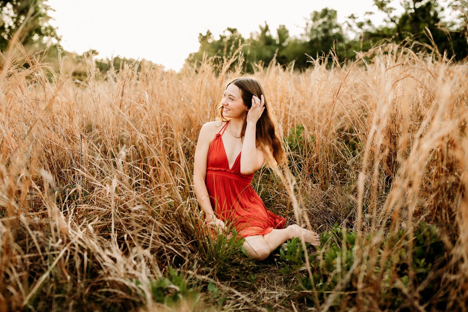 A figure in a flowing red dress walks through tall golden grass at sunset creating a dreamy outdoor portrait scene.