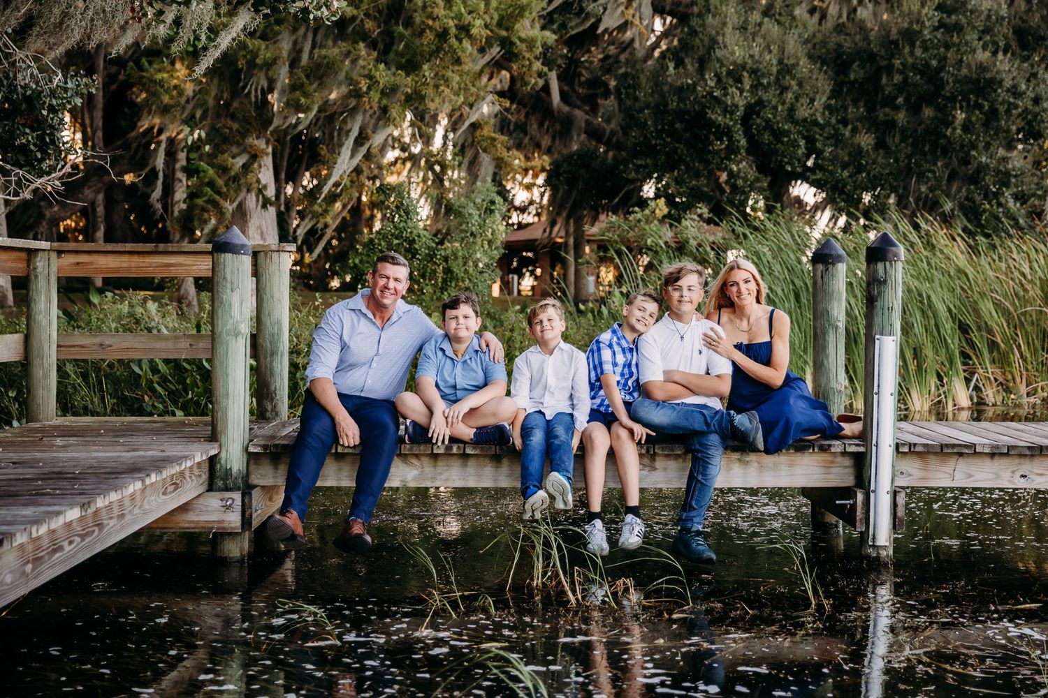 Family in coordinating blue outfits sits together on wooden dock over tranquil pond surrounded by lush greenery.