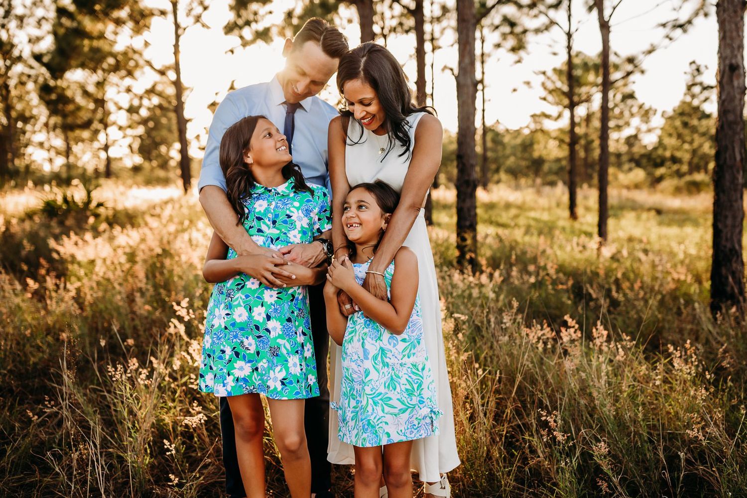 Family wearing coordinated blue outfits embraces while walking through a sunlit forest at golden hour.