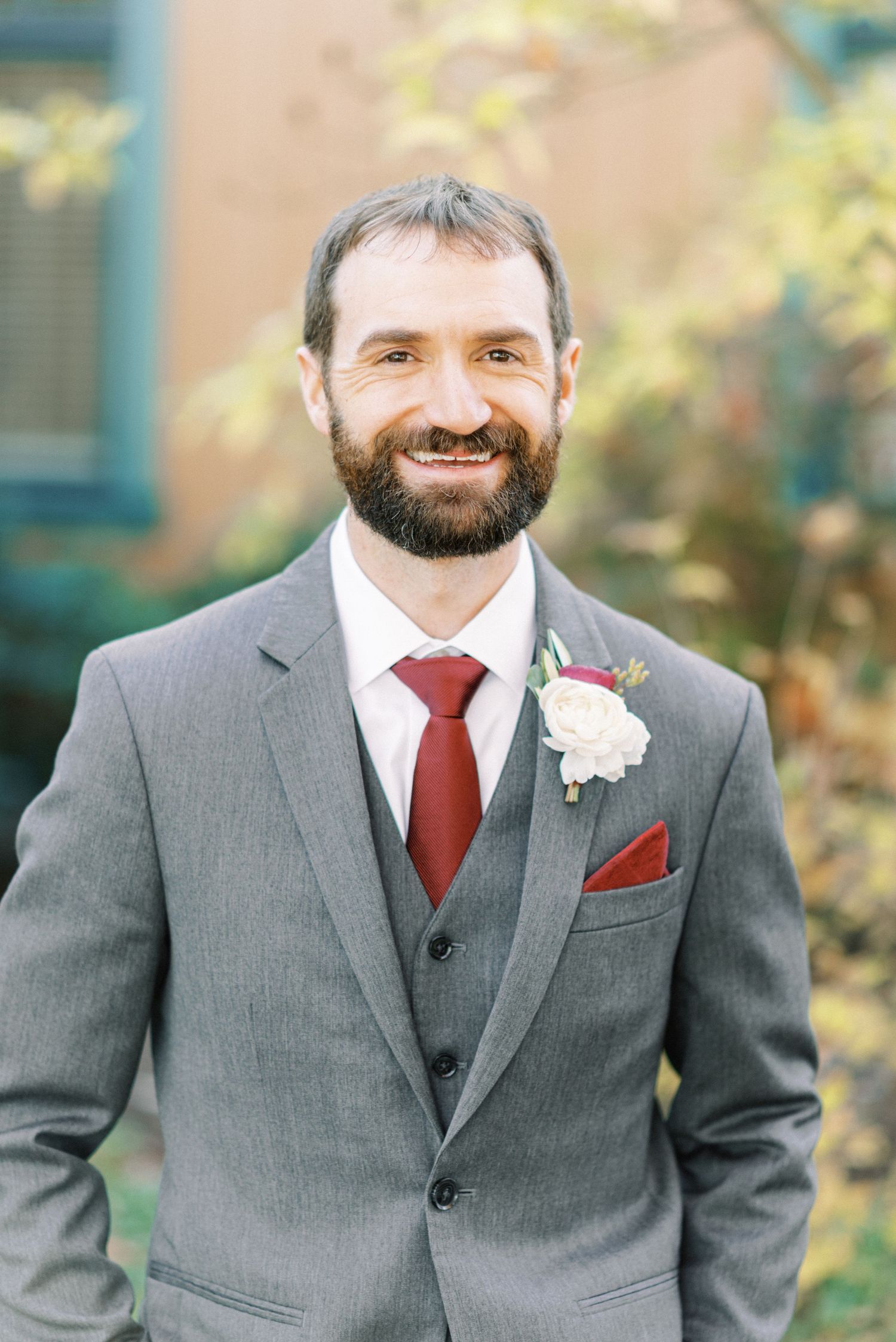 Man in a gray suit with a red tie and boutonniere smiles warmly. Background is softly blurred with fall foliage.